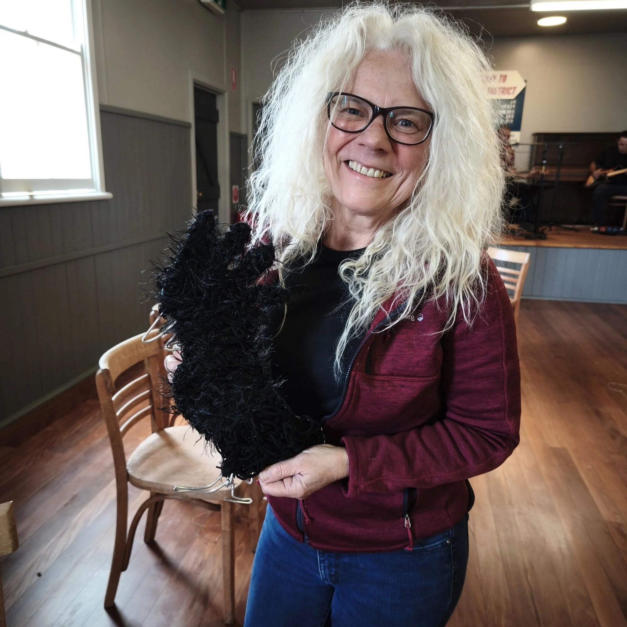 A cheerful woman with long, curly white hair wearing glasses, a burgundy jacket, and a black shirt, holding a black fluffy cockatoo scul, standing in a room with wooden floors and chairs in the background, and a musician playing guitar in the corner.