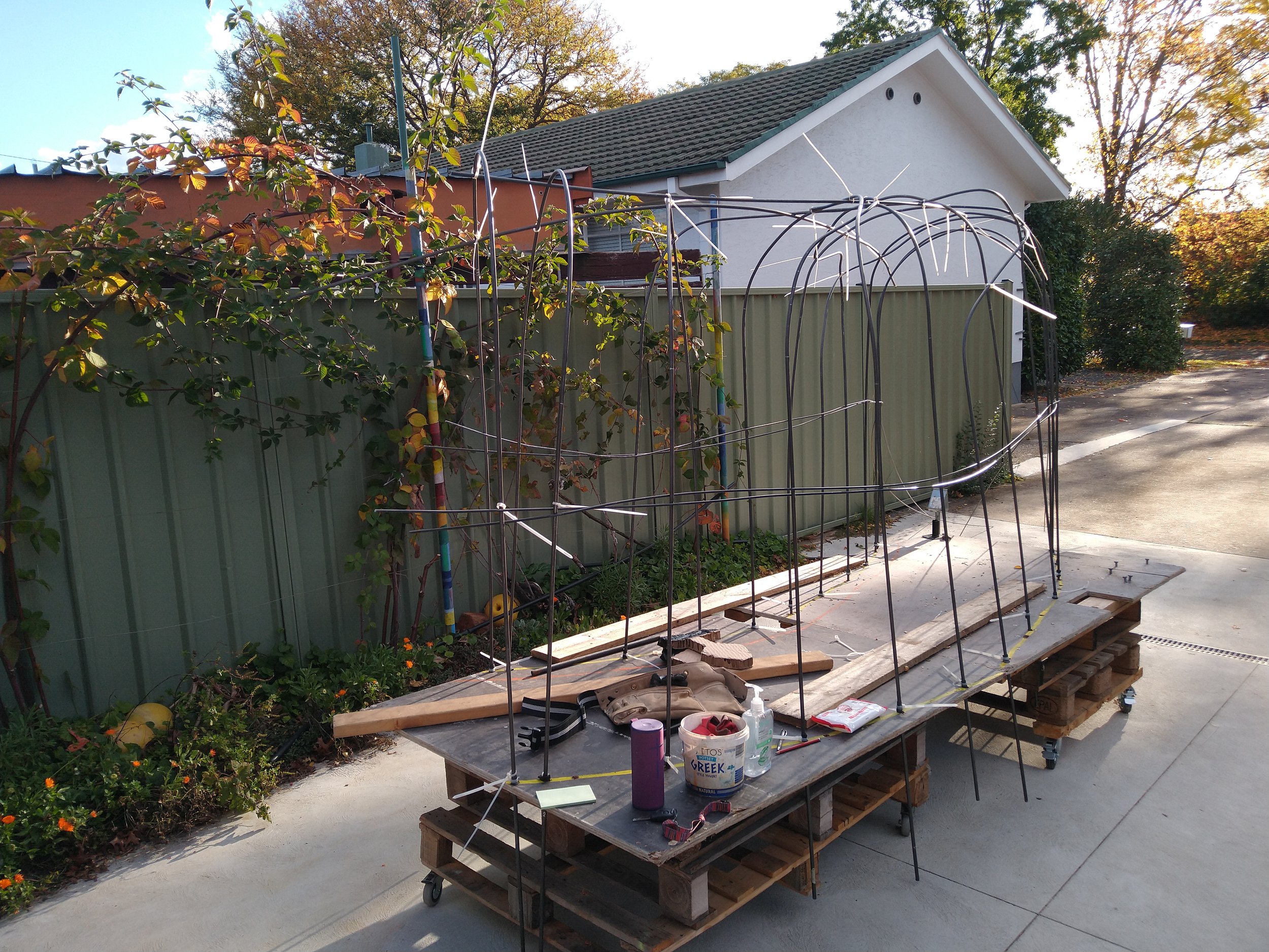 Outdoor workspace with pallets, tools, and materials as a maquette for the fibre sculpture 'The Bowery', set on a patio next to a house with a green fence and trees.