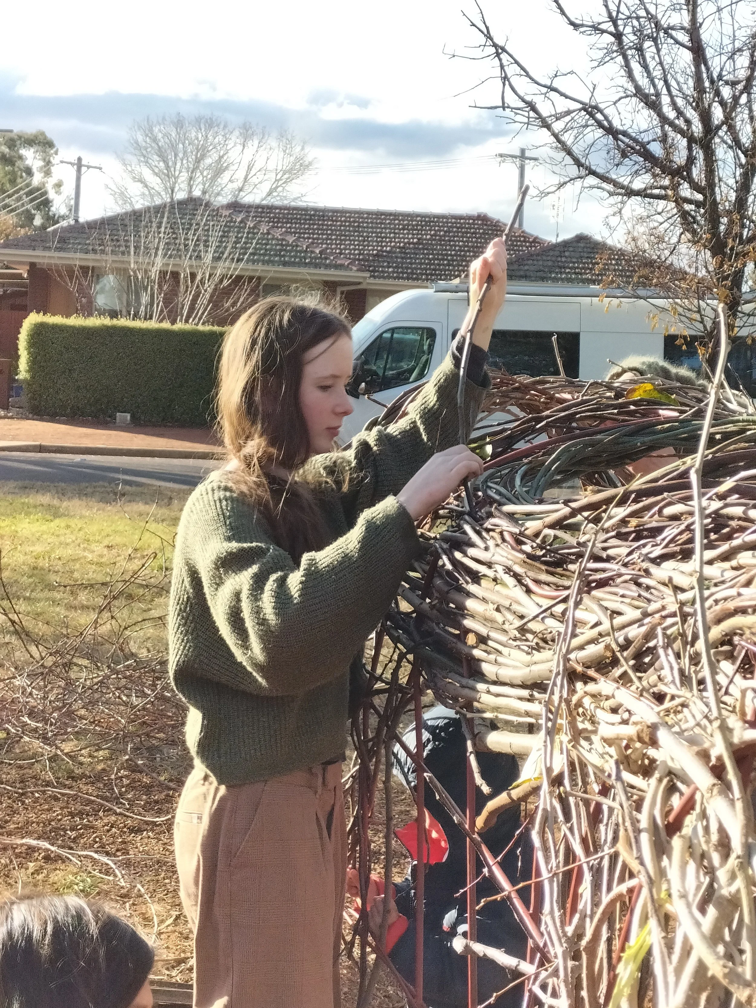 A young woman with long brown hair in a plaid shirt and beige pants is stringing or weaving twigs or branches in an outdoor setting with trees, a house, and a white van in the background.