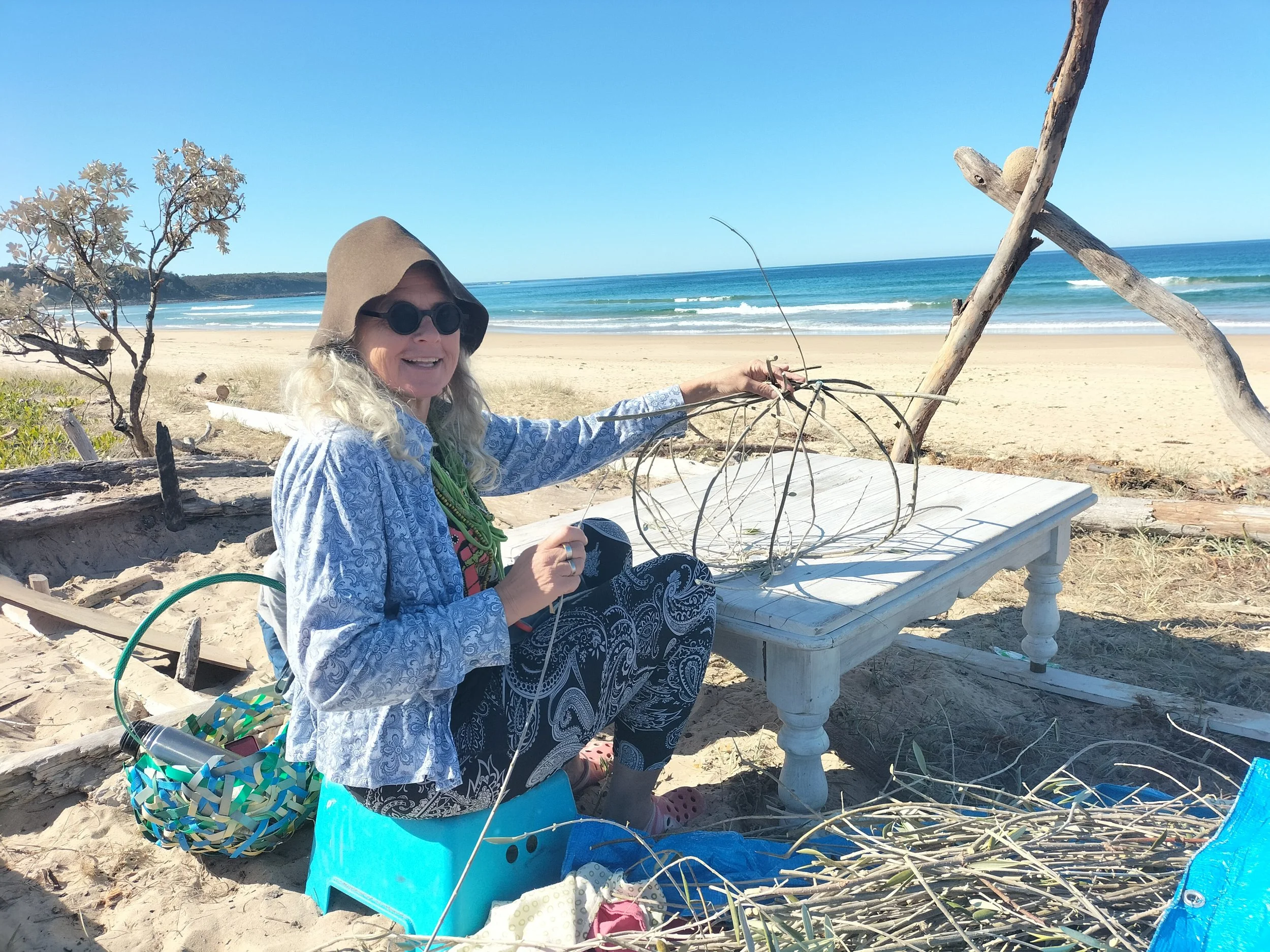 A woman with long blonde hair, wearing a hooded shirt and sunglasses, smiling while sitting on a bench on a sandy beach. She is holding a pile of thin branches. There is a colorful basket and some decorations around her, with the ocean, blue sky, and some trees in the background.