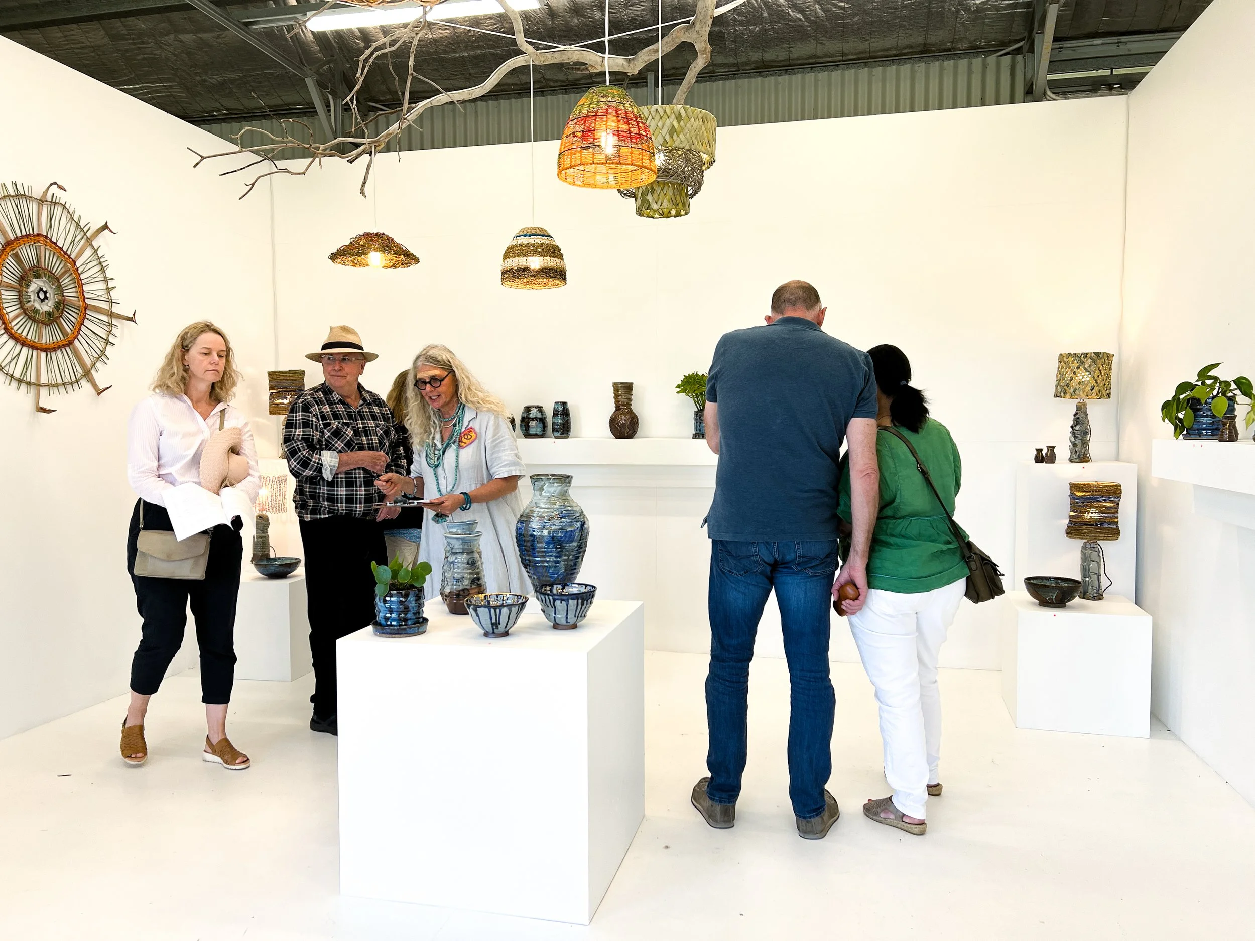 People browsing and admiring pottery and ceramic artworks in an art gallery or store, with various vases, bowls, and sculptures on white display tables and shelves.
