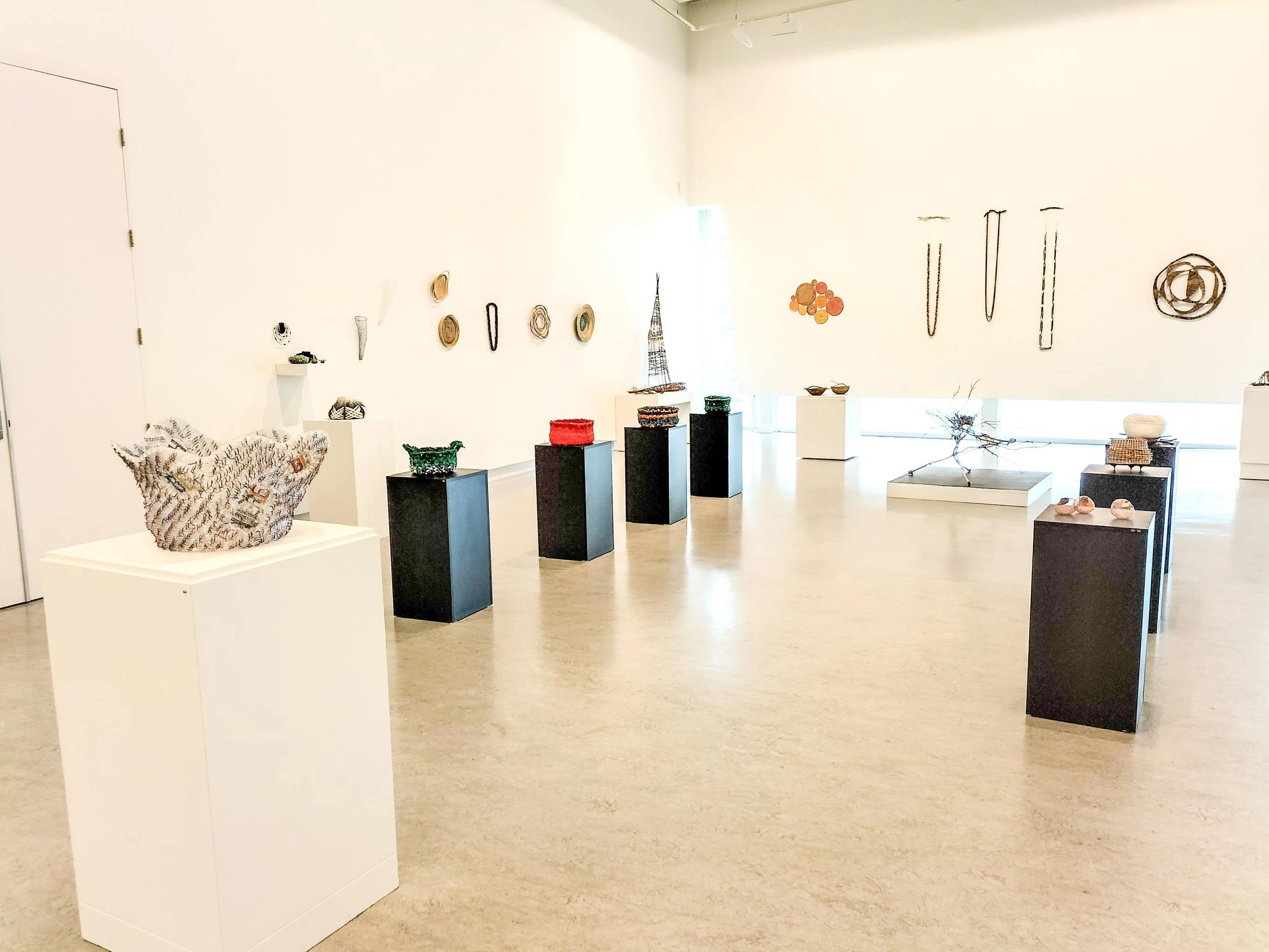 Art gallery with various baskets and sculptures displayed on white pedestals and black stands, with a large white wall holding woven art pieces.