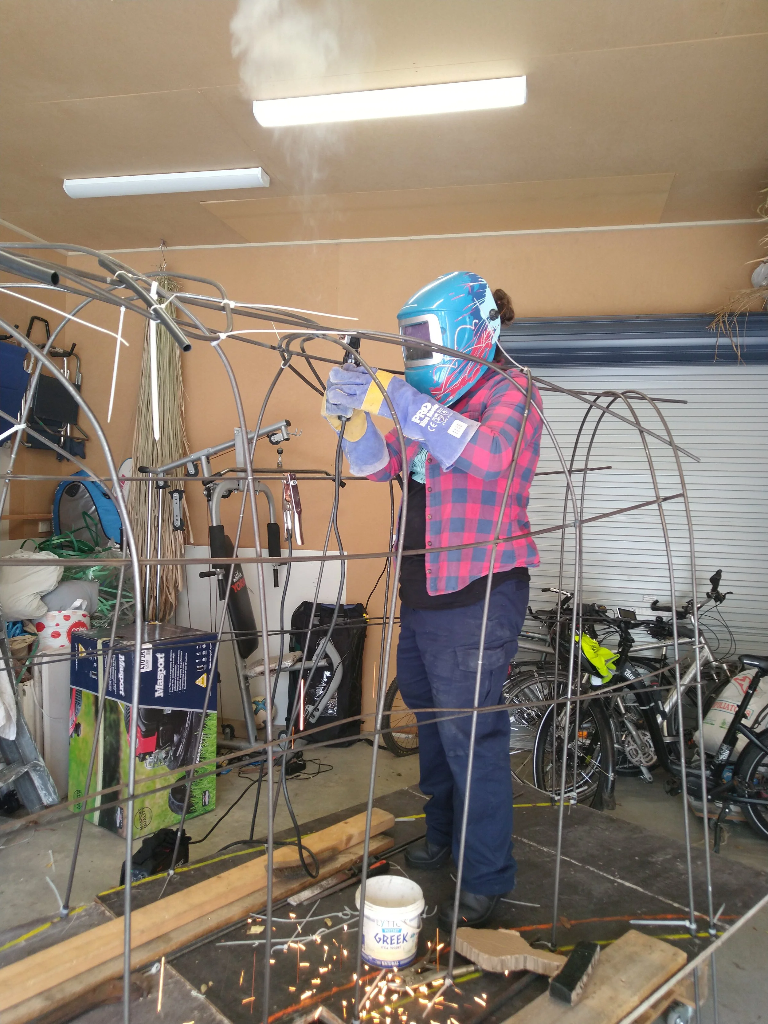 Person welding a metal frame in a workshop, wearing a blue welding helmet, gloves, plaid shirt, and jeans, with sparks flying from the welding process.