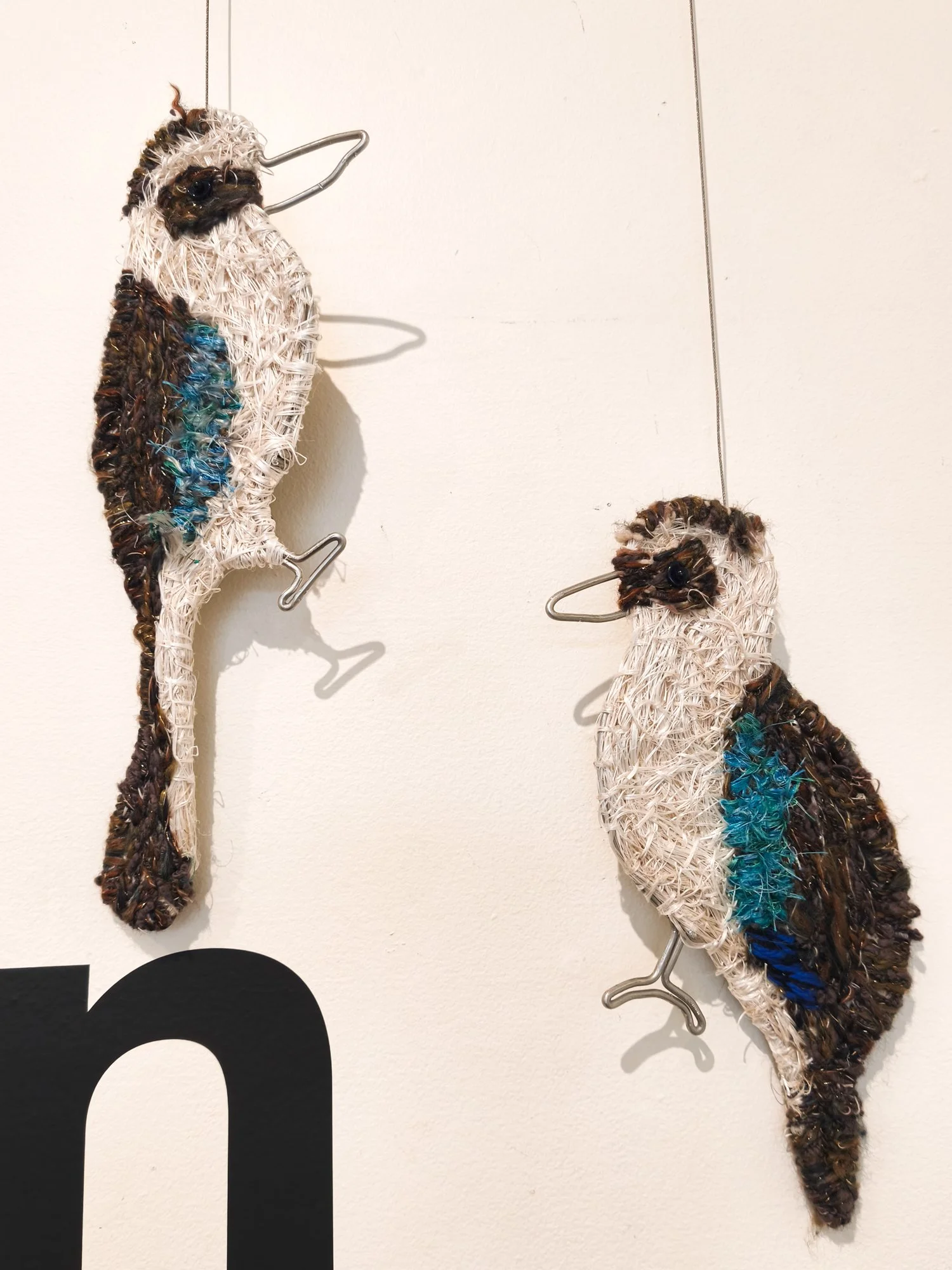 Two decorative fabric and wire kookaburra wall sculptures with wire hooks on a white wall, with a black letter or shape partially visible at the bottom.