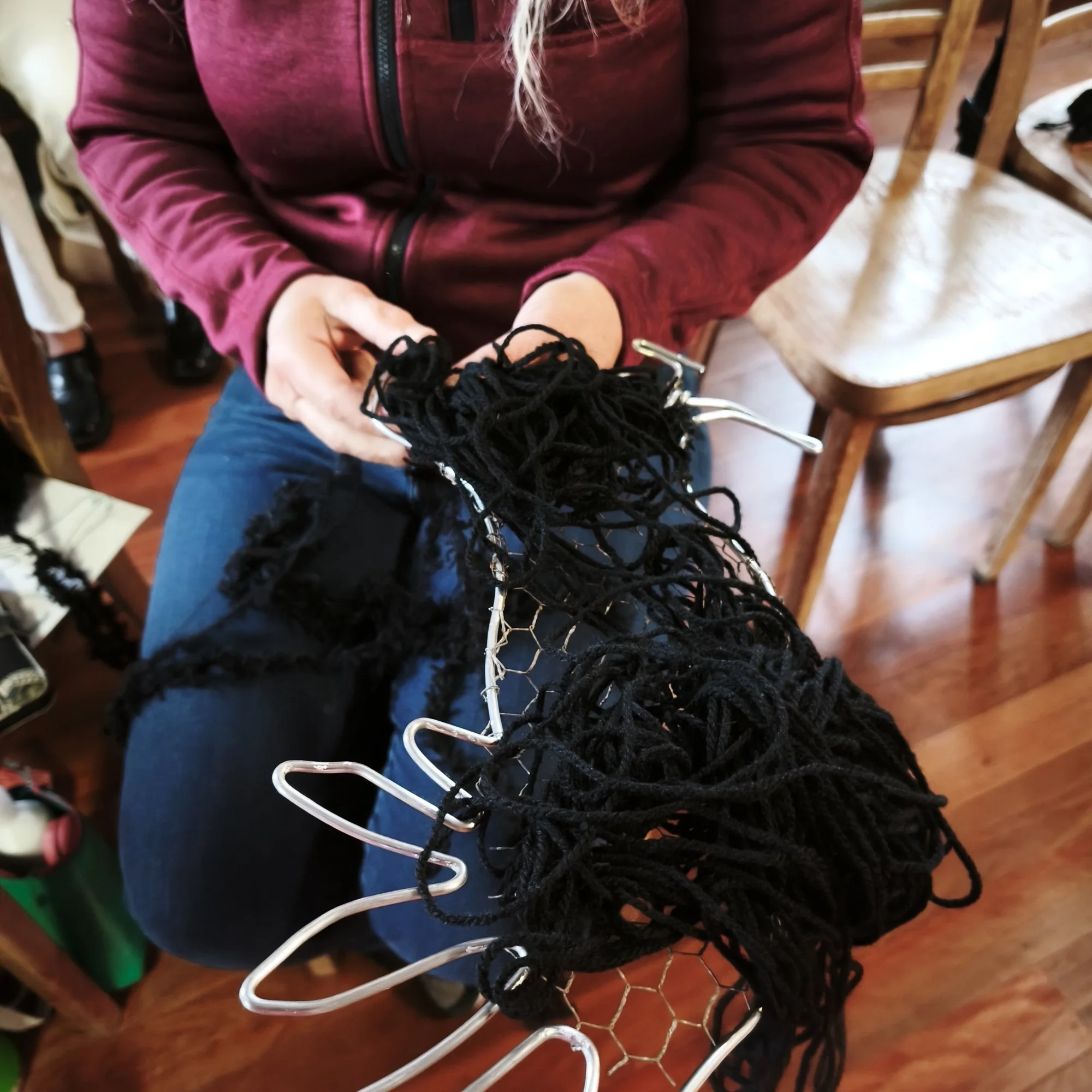 A person wearing a burgundy zip-up jacket holding black yarn and a white crochet hook, with several chairs and various items in a room.