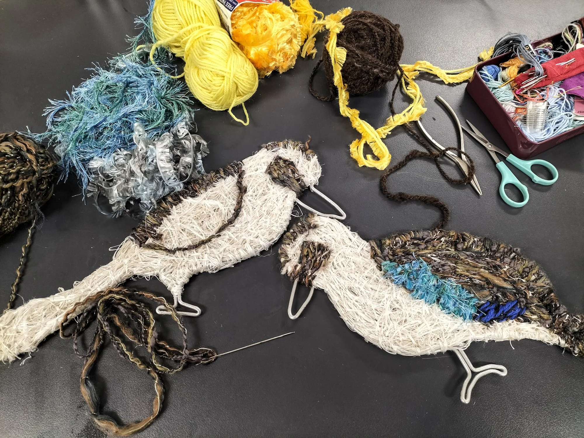Colourful yarns and threads used for fibre sculpture-making, with scissors and sewing tools on a black surface. 2 finished kookaburra sculptures are also in the image.