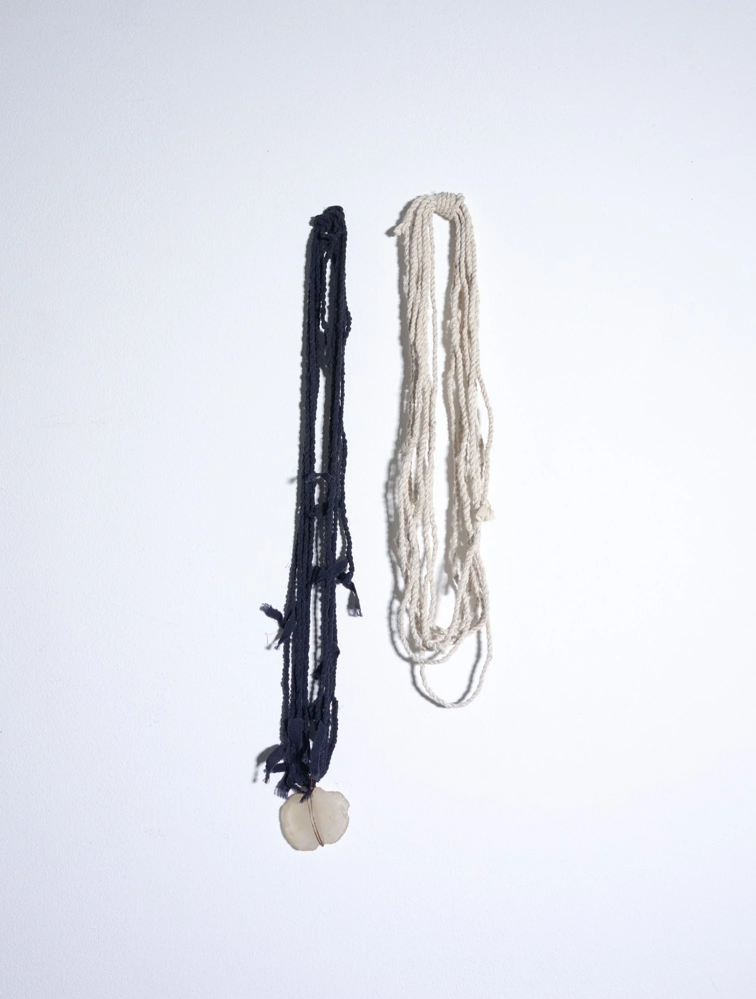 Two necklaces hanging against a white background, one dark blue and one cream-colored, with a shell pendant at the bottom.
