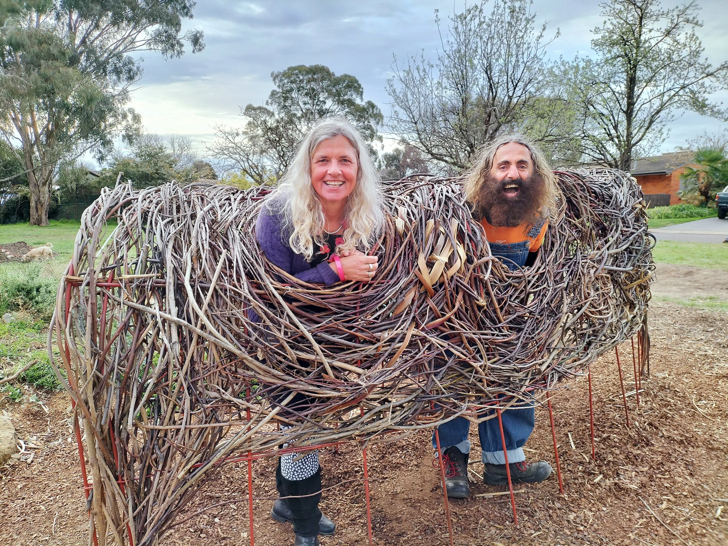 Two people with faces smiling are inside a large structure made of intertwined twigs and branches, resembling a nest or a sculpture, outdoors in a park with trees and cloudy sky in the background.