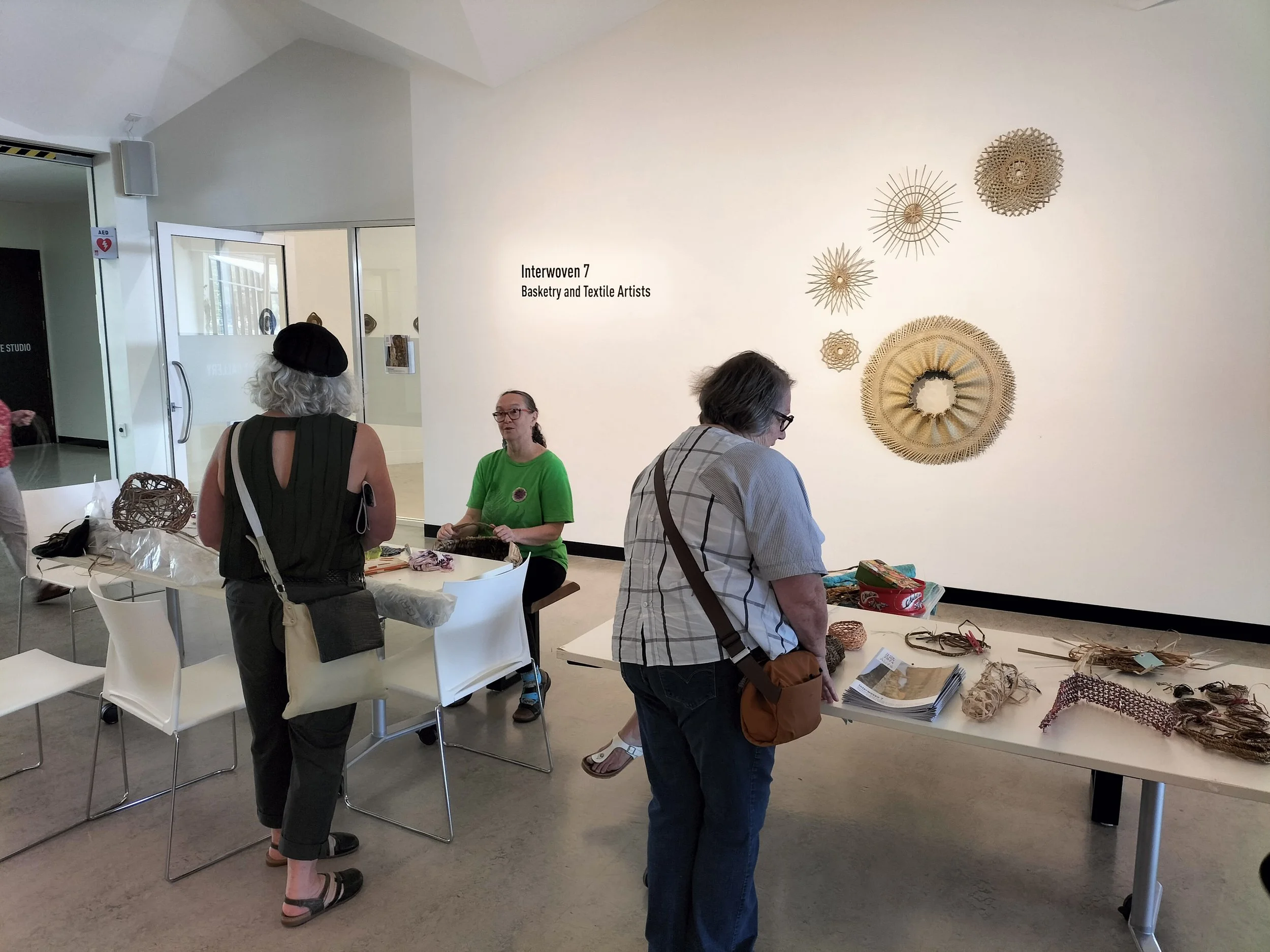 People browsing straw basketry and textile art pieces at an art exhibition. The display includes woven baskets and textile items on tables and wall art featuring straw patterns. The setting appears to be an indoor gallery space with attendees observing the artwork.