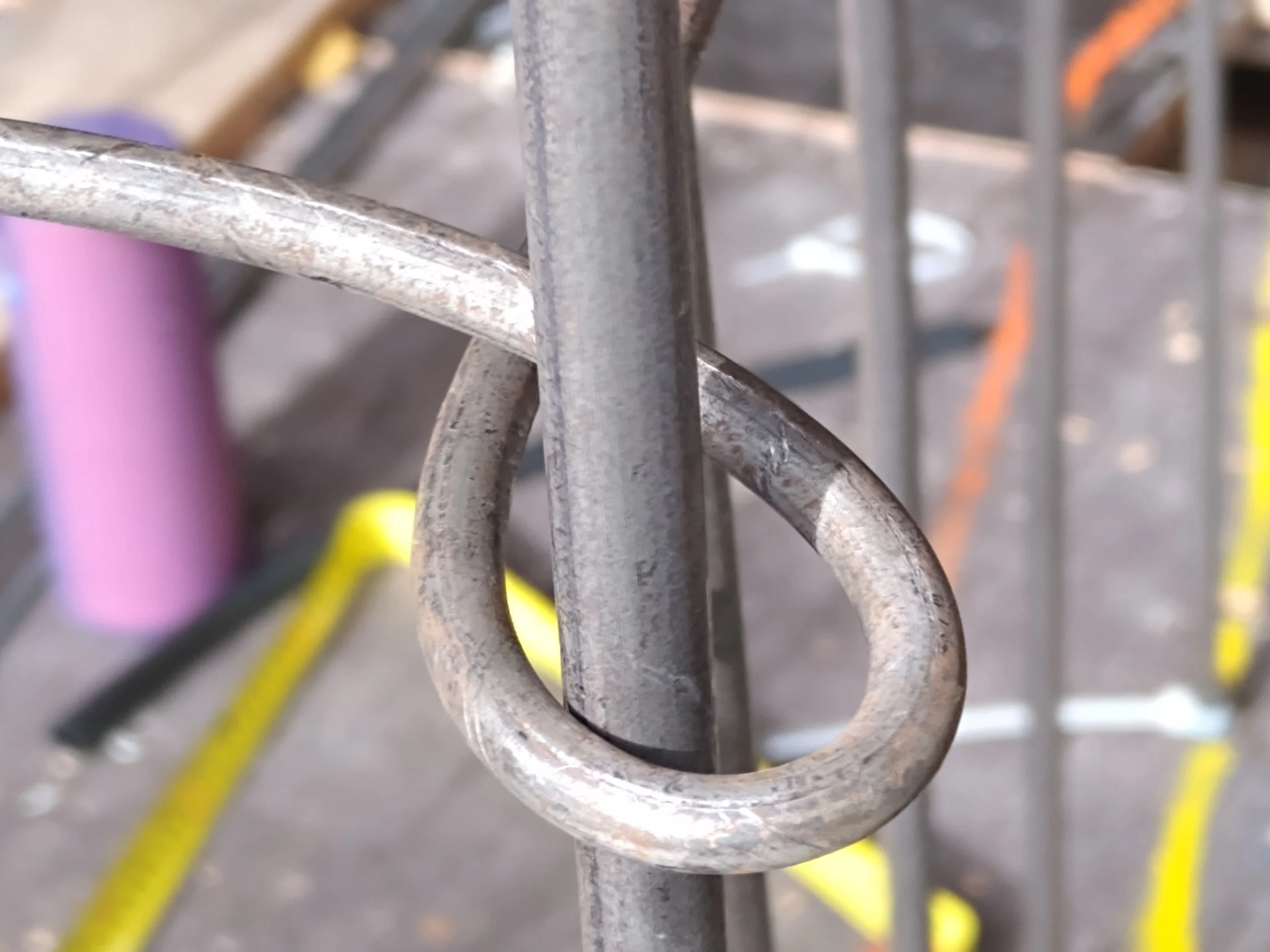 Close-up of a metal coil spring, one of the loops of the fibre sculpture frame of 'The Bowery', with a blurred background that includes pink, yellow, and orange objects.
