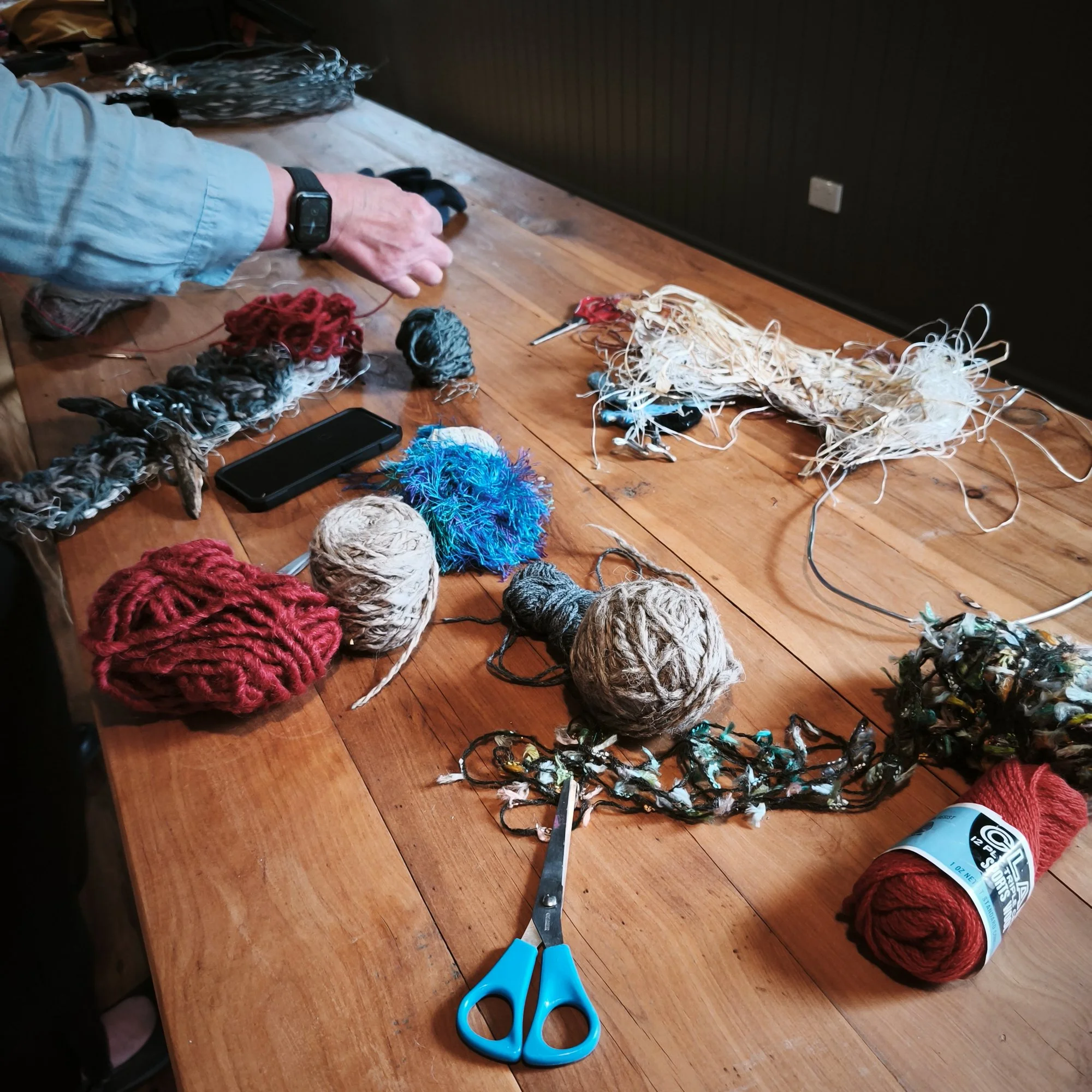 A wooden table with colorful yarn balls, scissors, a smartphone, and a craft glue bottle, in a workspace with a dark wall in the background.