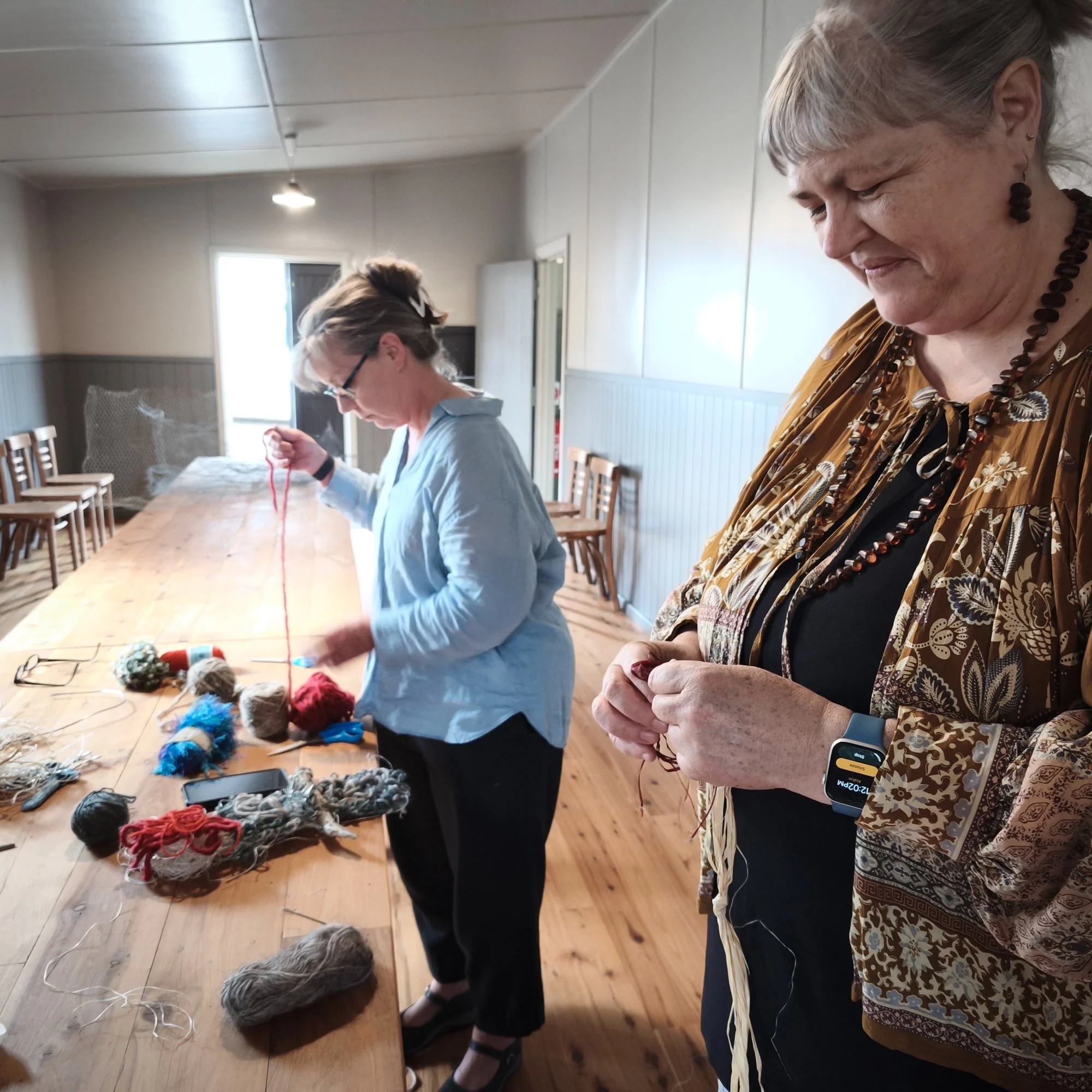 Two women in a room standing at a long wooden table with yarn and knitting supplies, one with earrings and colourful blouse, the other with glasses and grey hair, engaged in craft activities.