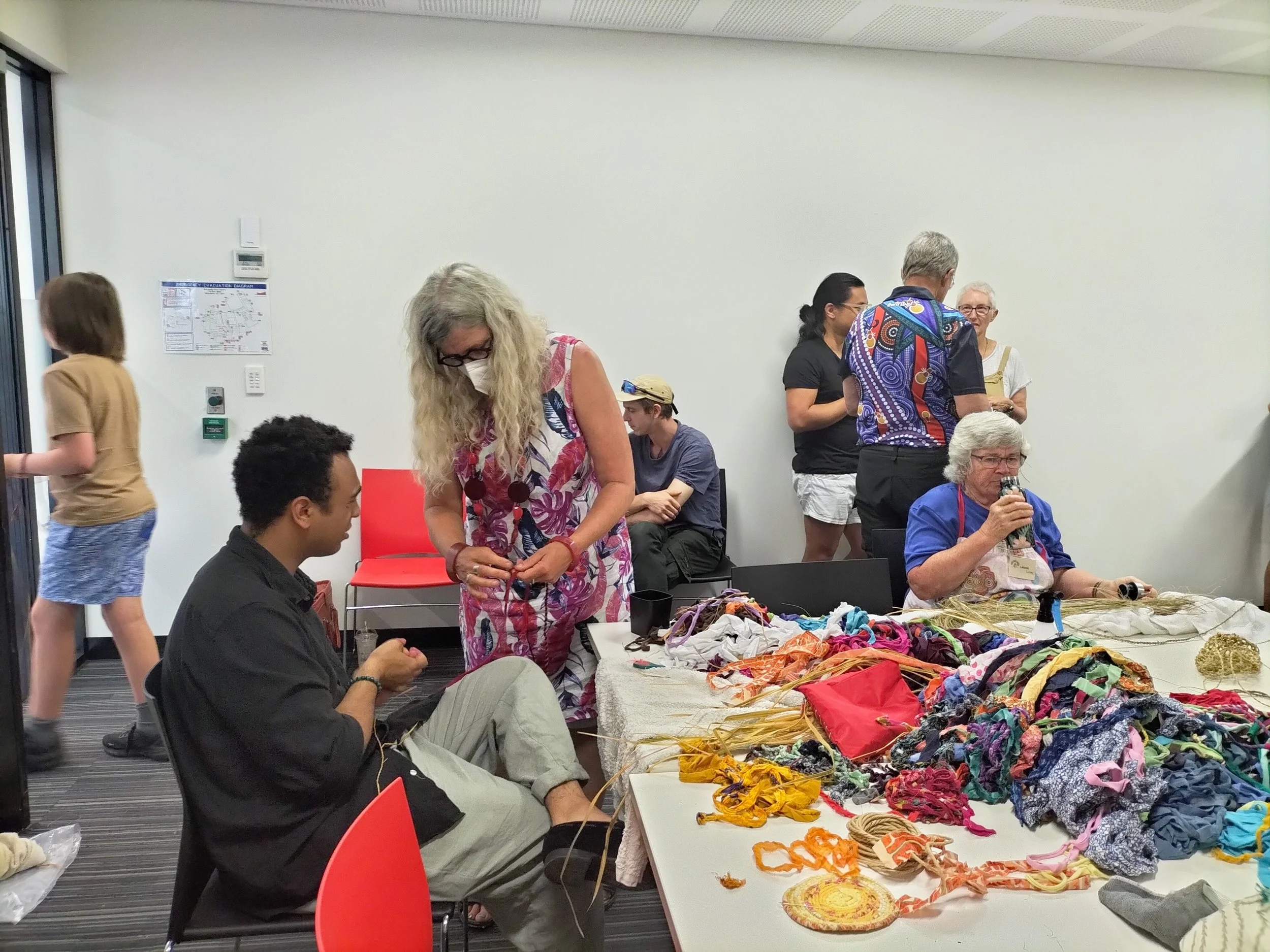 People gathered around a table displaying colourful fabric and craft items in a room with white walls and grey carpet. A weaving workshop at Belconnen Arts Centre
