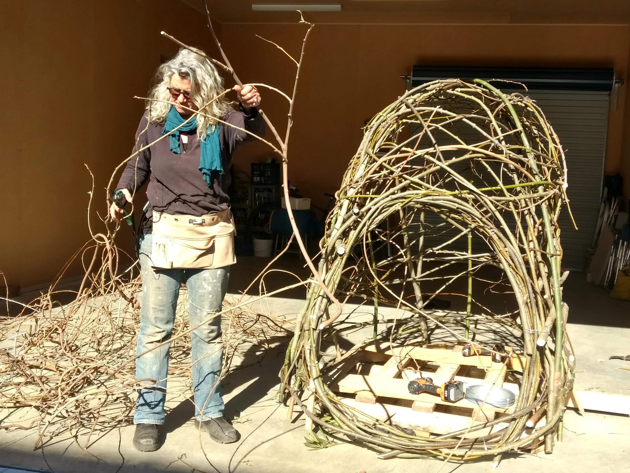 A woman with curly gray hair, wearing glasses, a dark jacket, and beige apron, stands among large, woven wicker structures made from intertwined branches, inside a workshop or garage with a closed rolling shutter in the background. She is holding a branch and working.