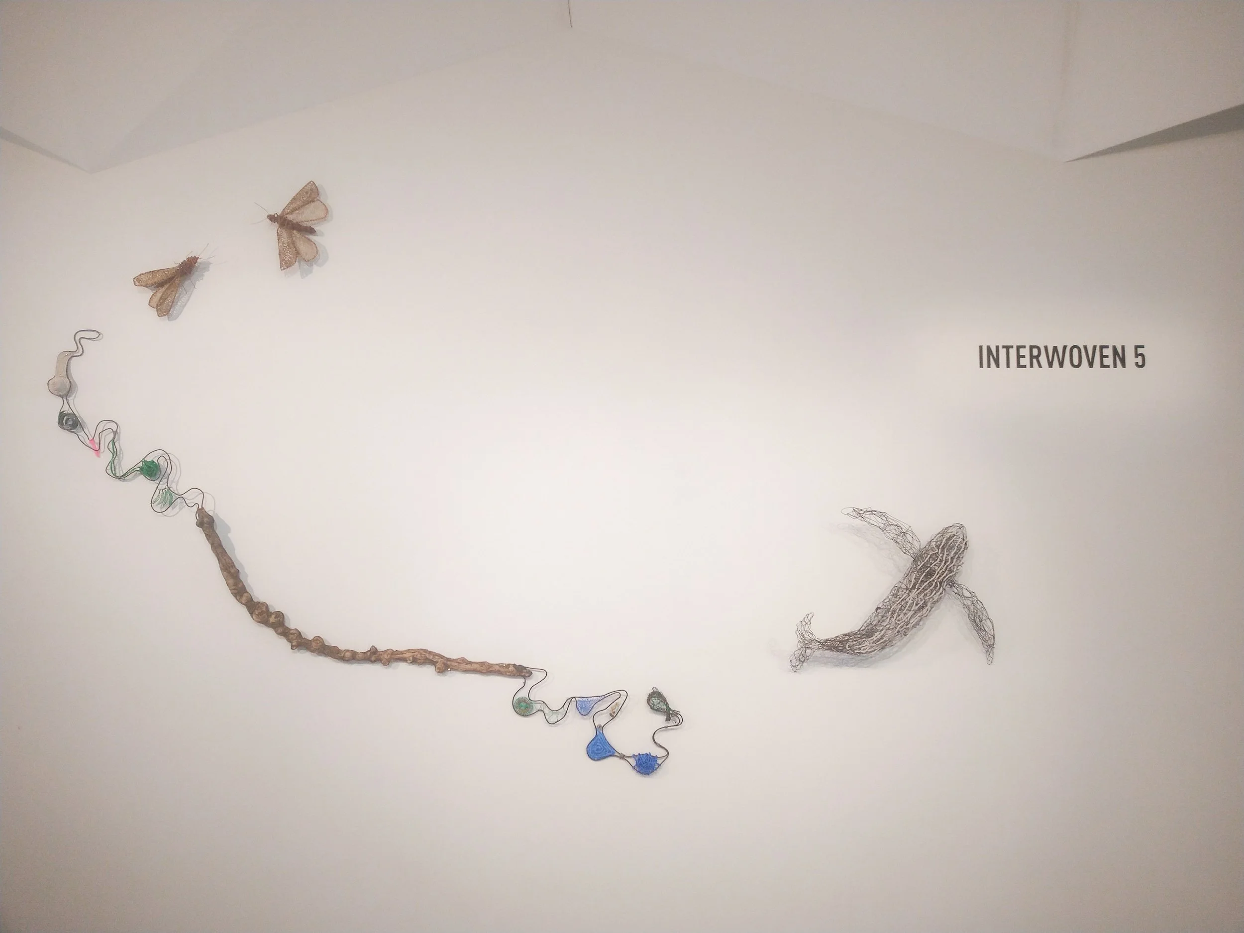 Decorative wall art with the label 'INTERWOVEN 5,' featuring fabric, wire, and paper elements resembling insects and abstract shapes.