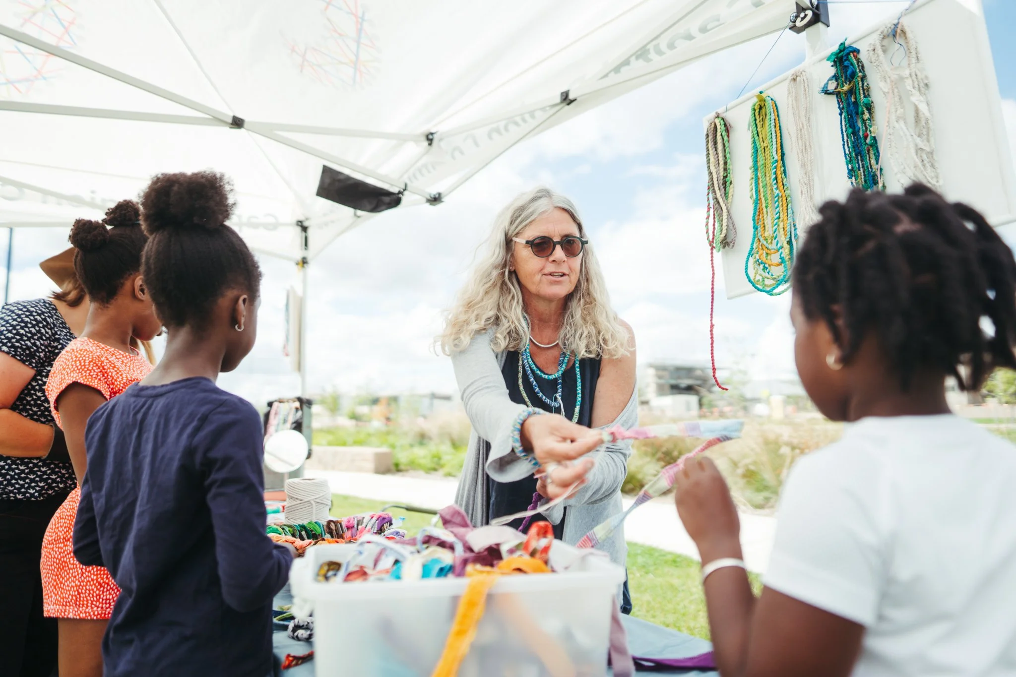 A woman is helping workshop participants to choose the required materials to make string jewellery at an outdoor event under a white canopy tent, with young girls talking to her.