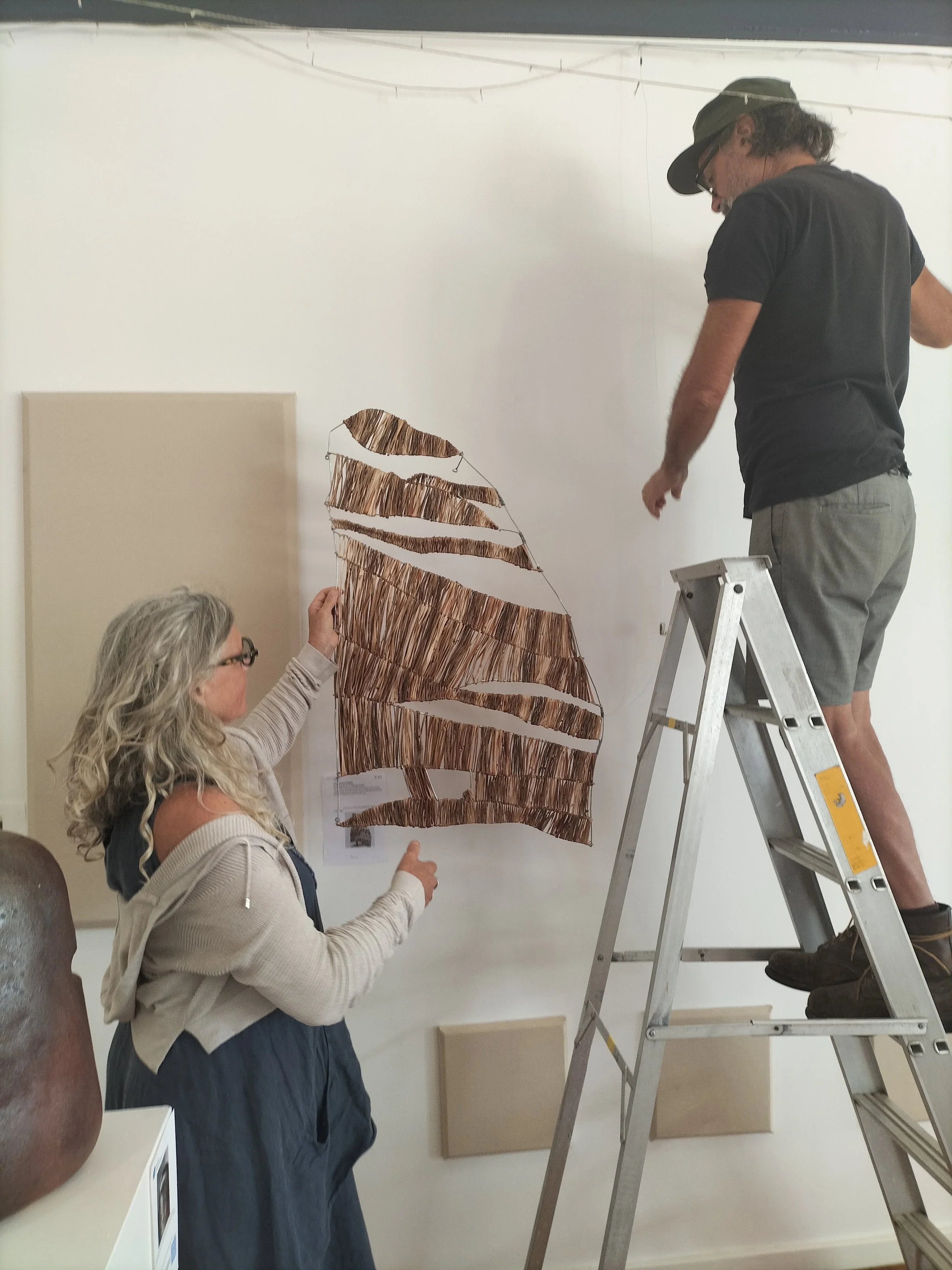 Two women and one man working together to hang a large, rust-colored, abstract sculpture made of metal strips on a white wall in an art gallery. The woman with gray hair and glasses is holding the sculpture, the other woman is assisting, and the man is standing on a ladder.