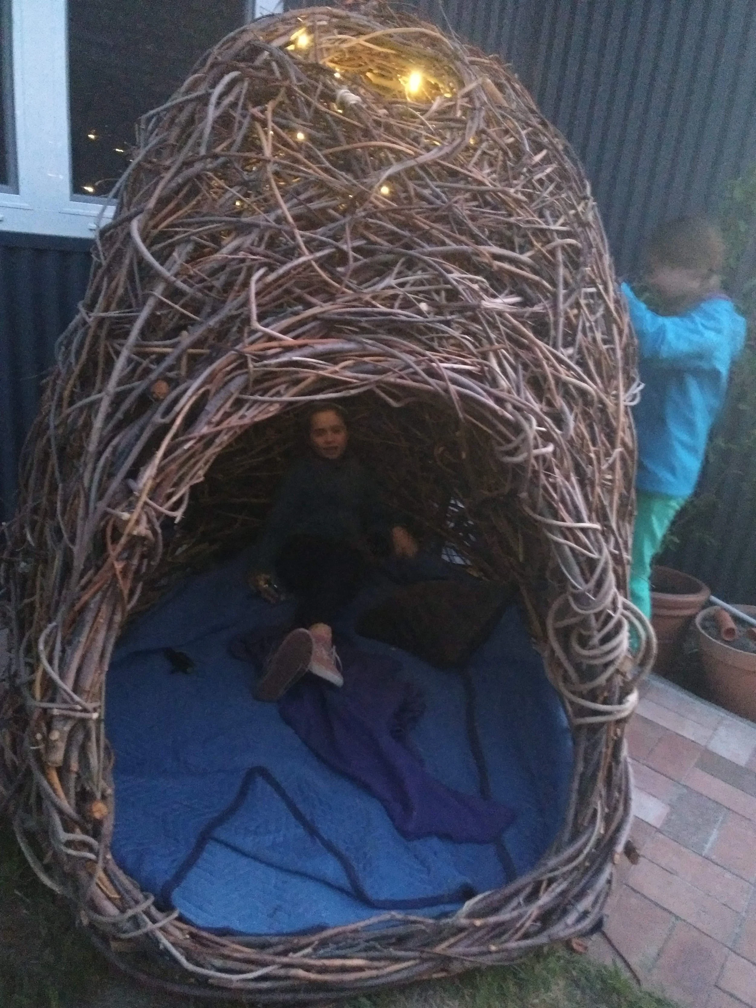 Child sitting inside a large woven fibre sculpture entitled 'nest' with a blue cushion, outdoors near a house at dusk.