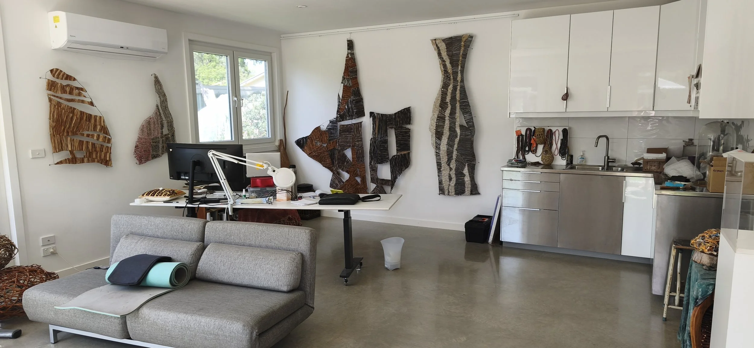 Living space with a desk, computer, and wall art made of woven material. Sofa with yoga mat, in front of kitchen with cabinets and sink. Various items on counter, floor, and furniture.