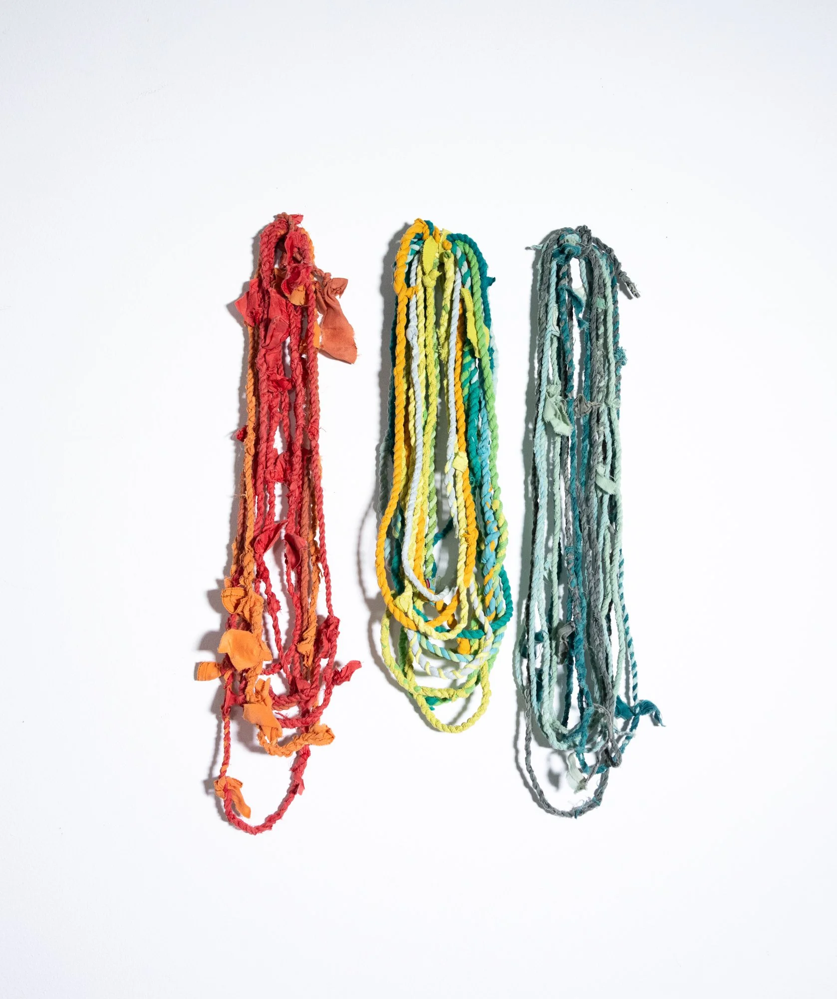Three colorful strands of torn and frayed fabric yarns hanging on a white background, from left to right in red, yellow, and gray colors.