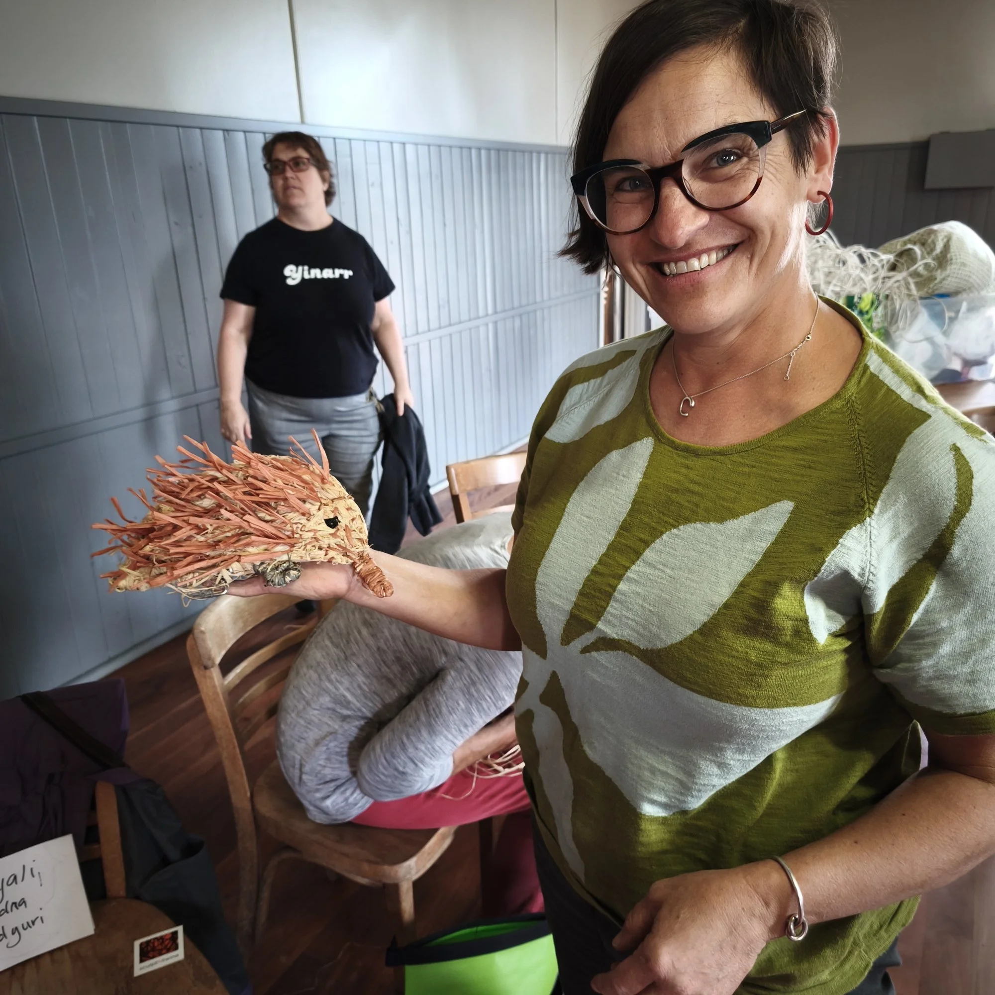 A woman with short dark hair, glasses, and earrings smiling while holding a fibre that resembles a gang gang bird. In the background, a person with glasses, wearing a black t-shirt, stands near a grey wall.