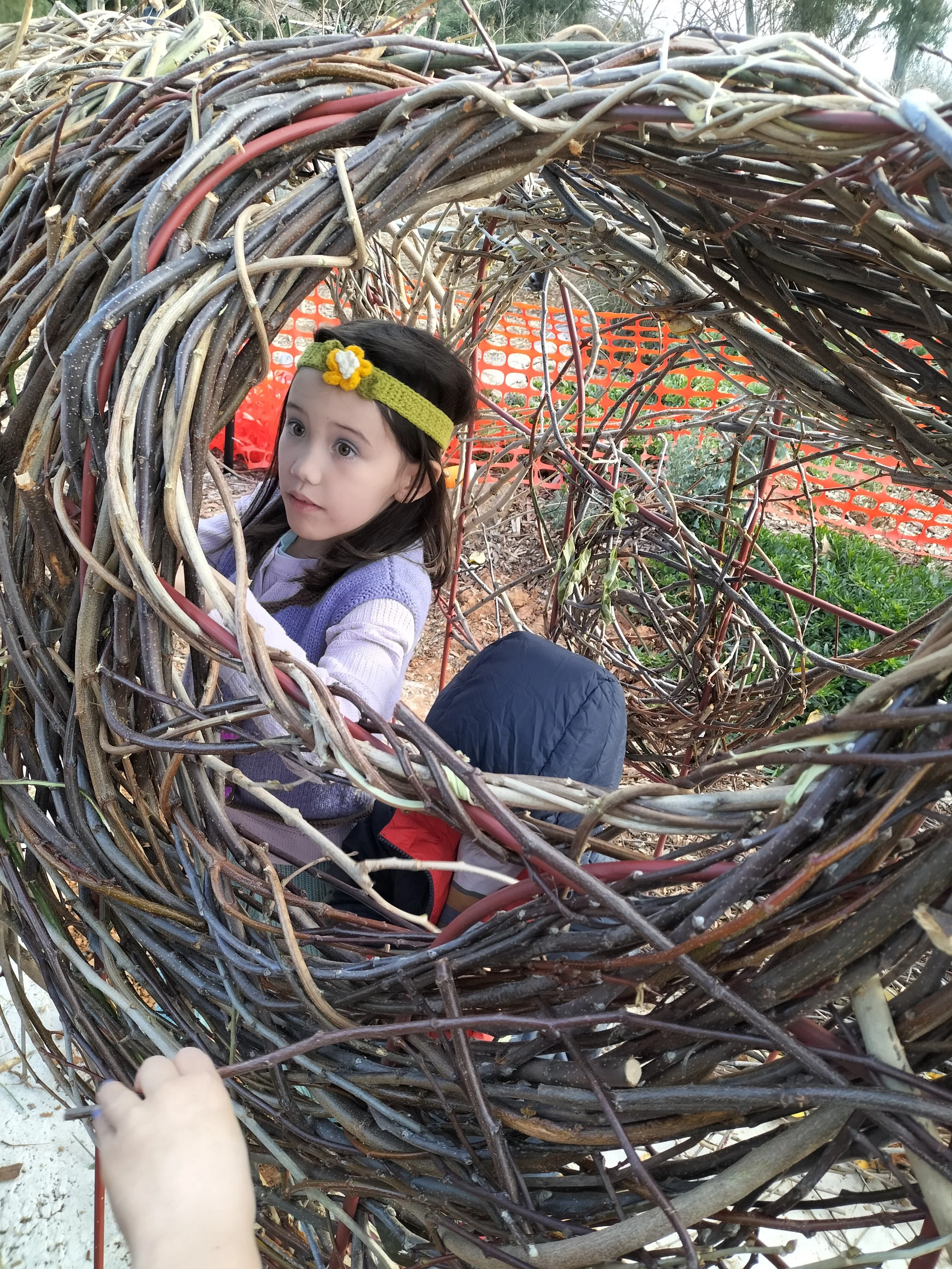 A young girl with dark hair and a yellow headband with a flower, looking out from inside a large nest made of intertwined sticks, with a person’s hand visible in the foreground.