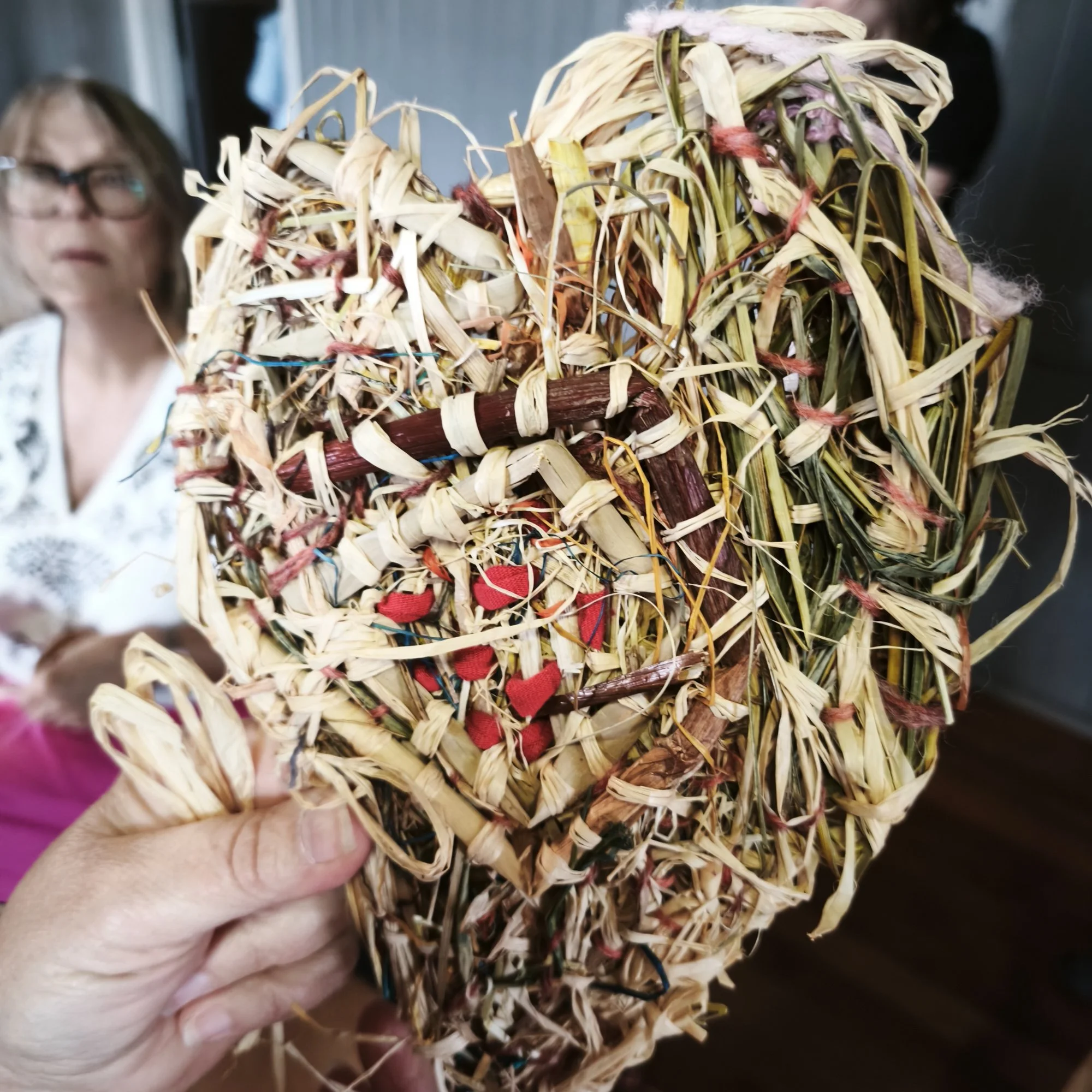 Close-up of a hand holding a small bundle of woven raffia and twine in the shape of a heart, with a blurred background including a woman wearing glasses.