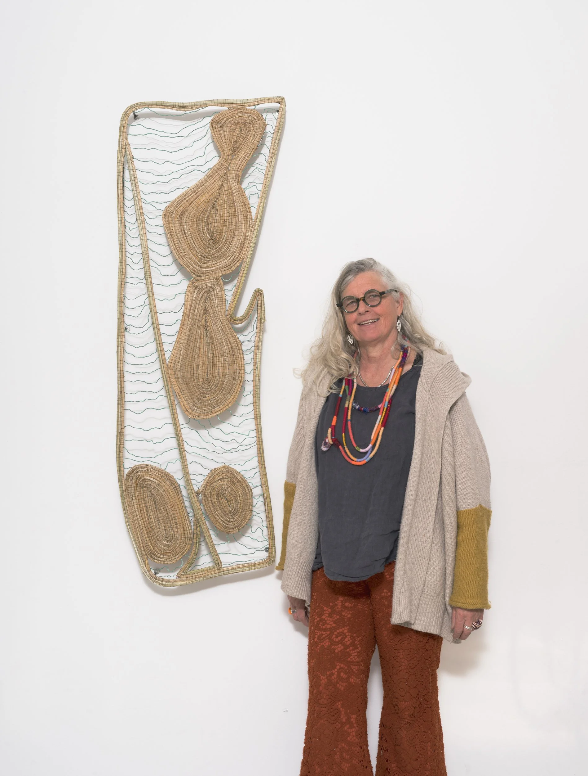 An older woman with long gray hair, wearing glasses, a multicolored necklace, a gray top, a beige cardigan with mustard yellow sleeves, and rust-colored lace pants, standing next to a woven art piece on a white wall.