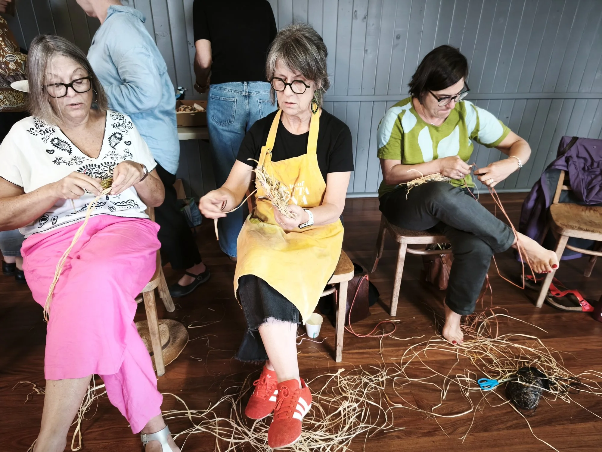 Three women sitting on chairs engaged in a craft activity involving raffia, with scattered craft materials and a light blue wooden wall in the background.