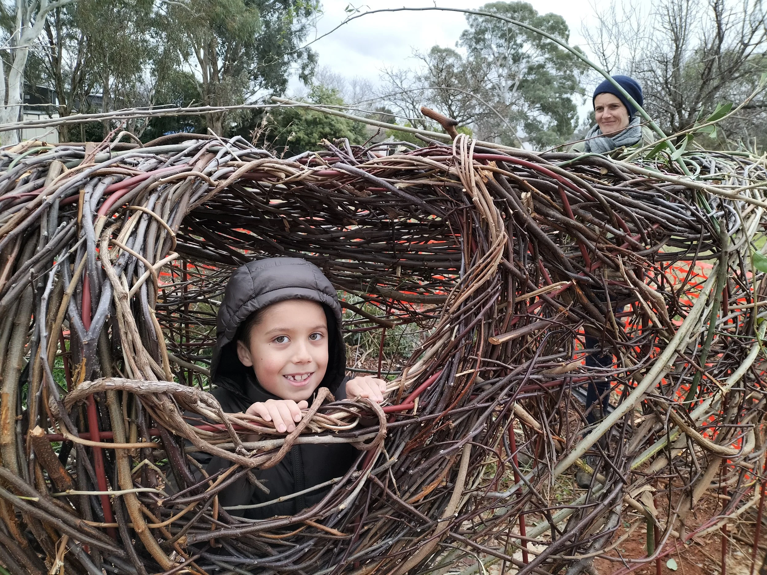 A boy with a black jacket and hood, smiling and holding onto the edge of a large, woven stick structure. Behind him is a woman with a scarf and beanie, smiling, standing among trees and outdoors.