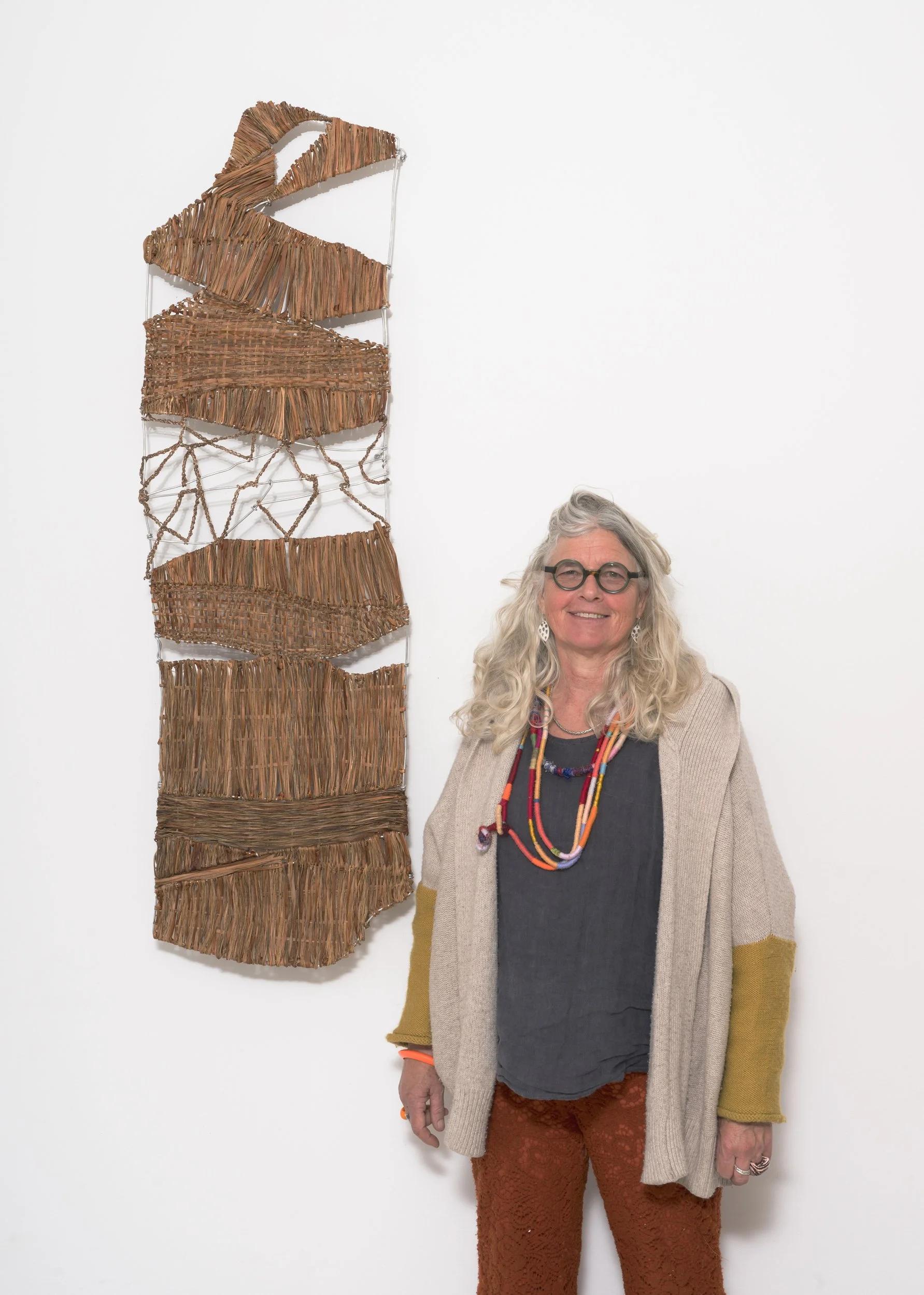 woman with long gray curly hair, glasses, wearing a beige and yellow cardigan, dark gray shirt, orange lace pants, standing next to a wall with woven artwork hanging on it.