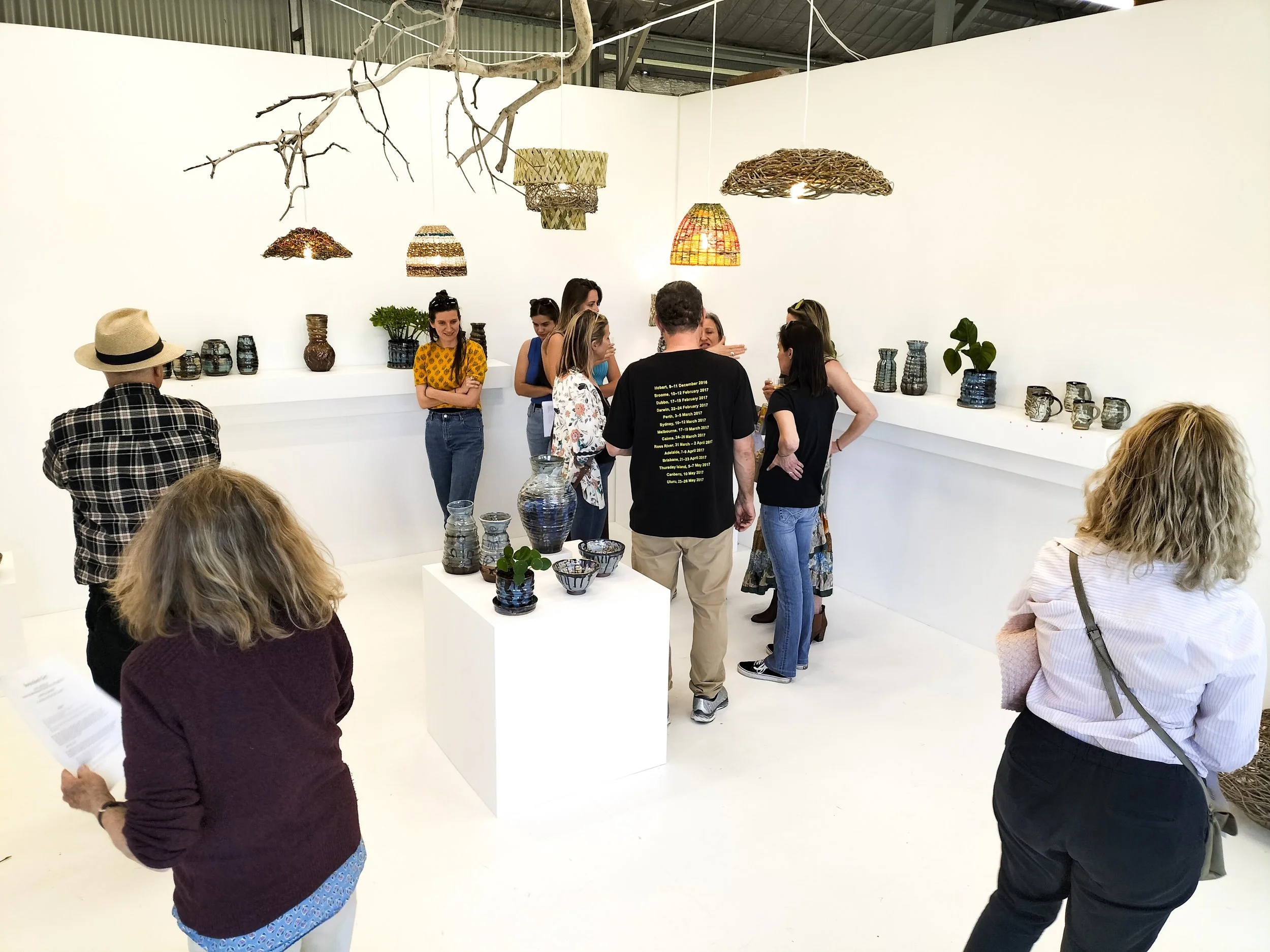 People gathered around art displays in an art gallery, examining pottery and ceramic pieces on white shelves, with hanging woven light fixtures and a white wall in the background.