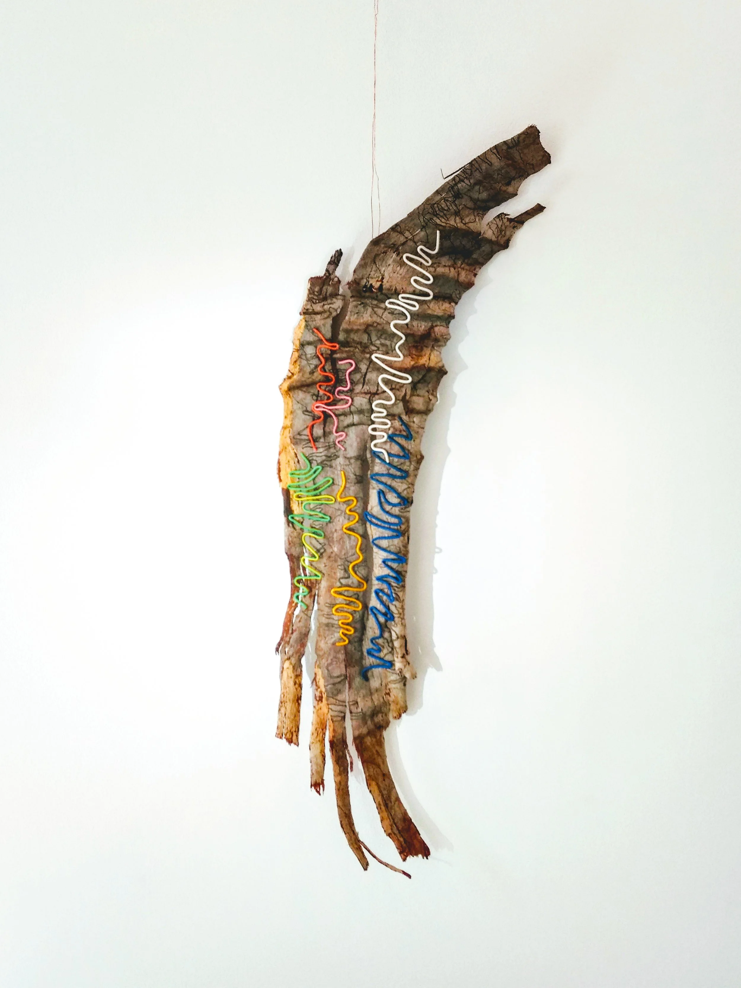 A piece of bark with colorful wire art attached, displaying various squiggly lines in red, pink, yellow, green, blue, and white, on a white background.