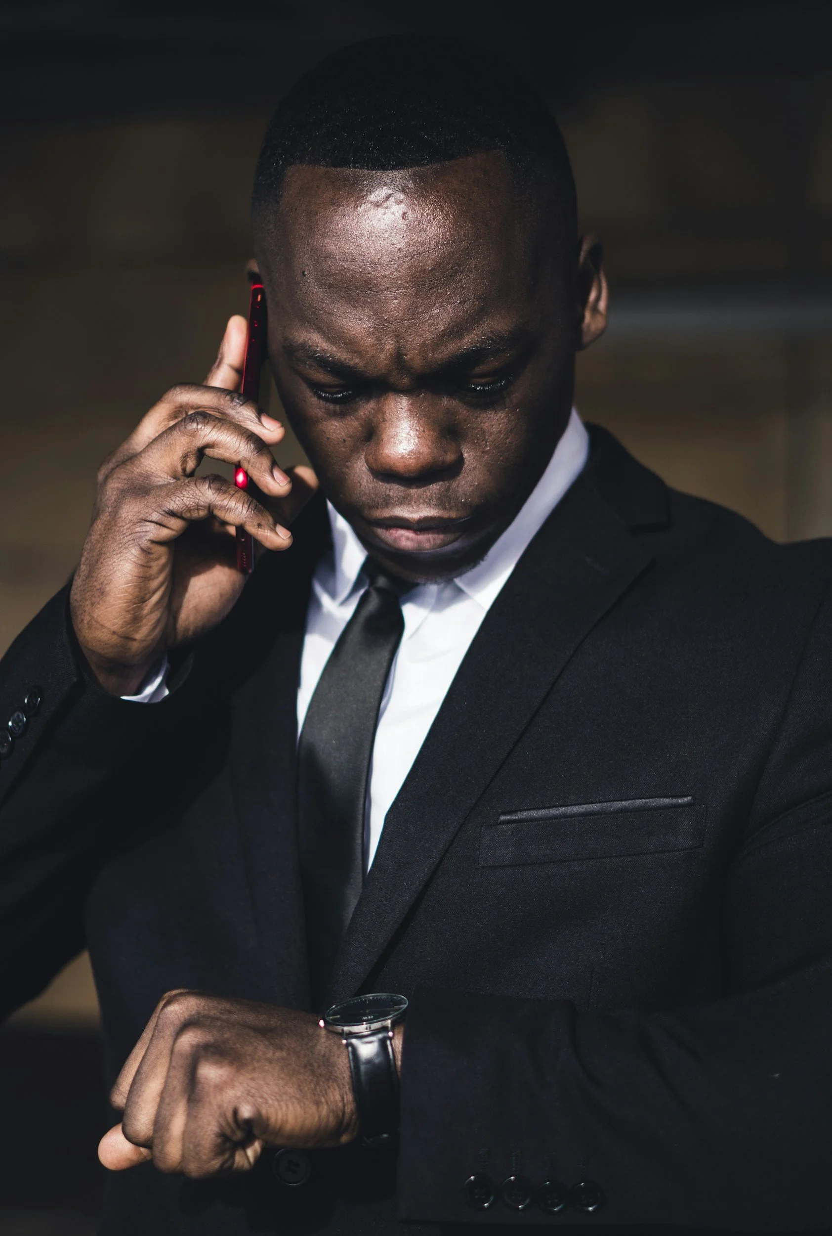 Un homme en costume noir blanc avec une montre vérifie l'heure et parle au téléphone portable, portant une montre noire, dans un environnement intérieur.