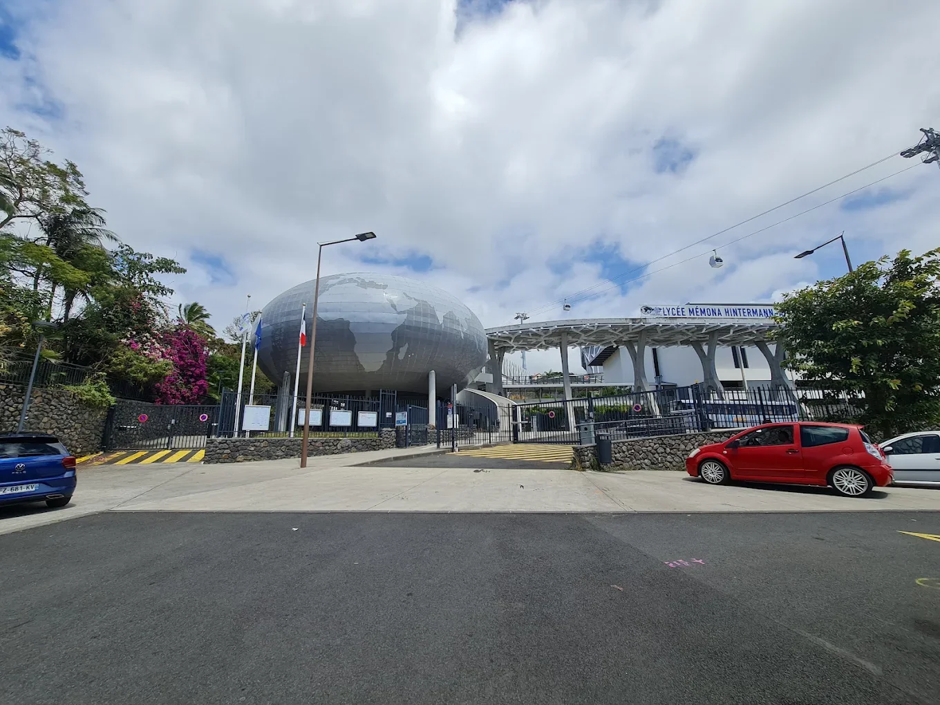 Le bâtiment du lycée Mé Mona Hintermann avec une structure moderne, un globe terrestre en facade, et quelques voitures garées devant.