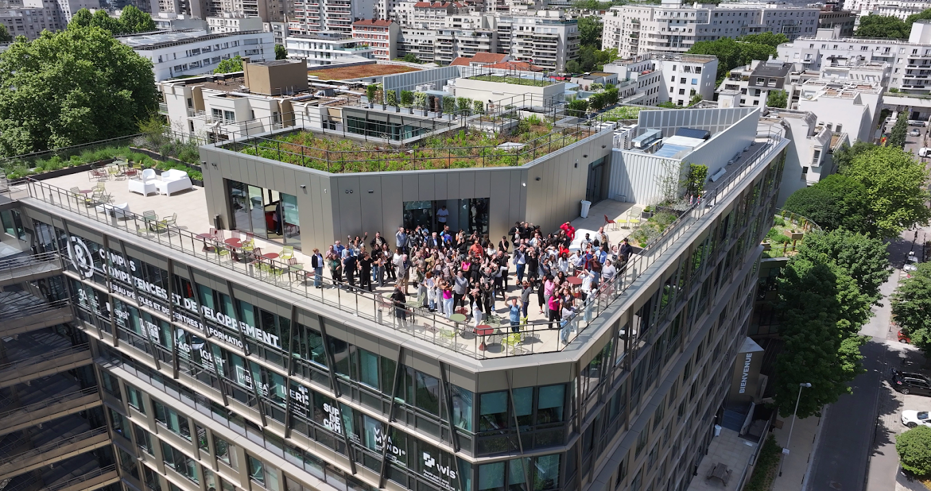 Une photo aérienne d'un bâtiment moderne avec une terrasse sur le toit où une foule de personnes se rassemble, entourée par un environnement urbain avec plusieurs bâtiments et arbres.