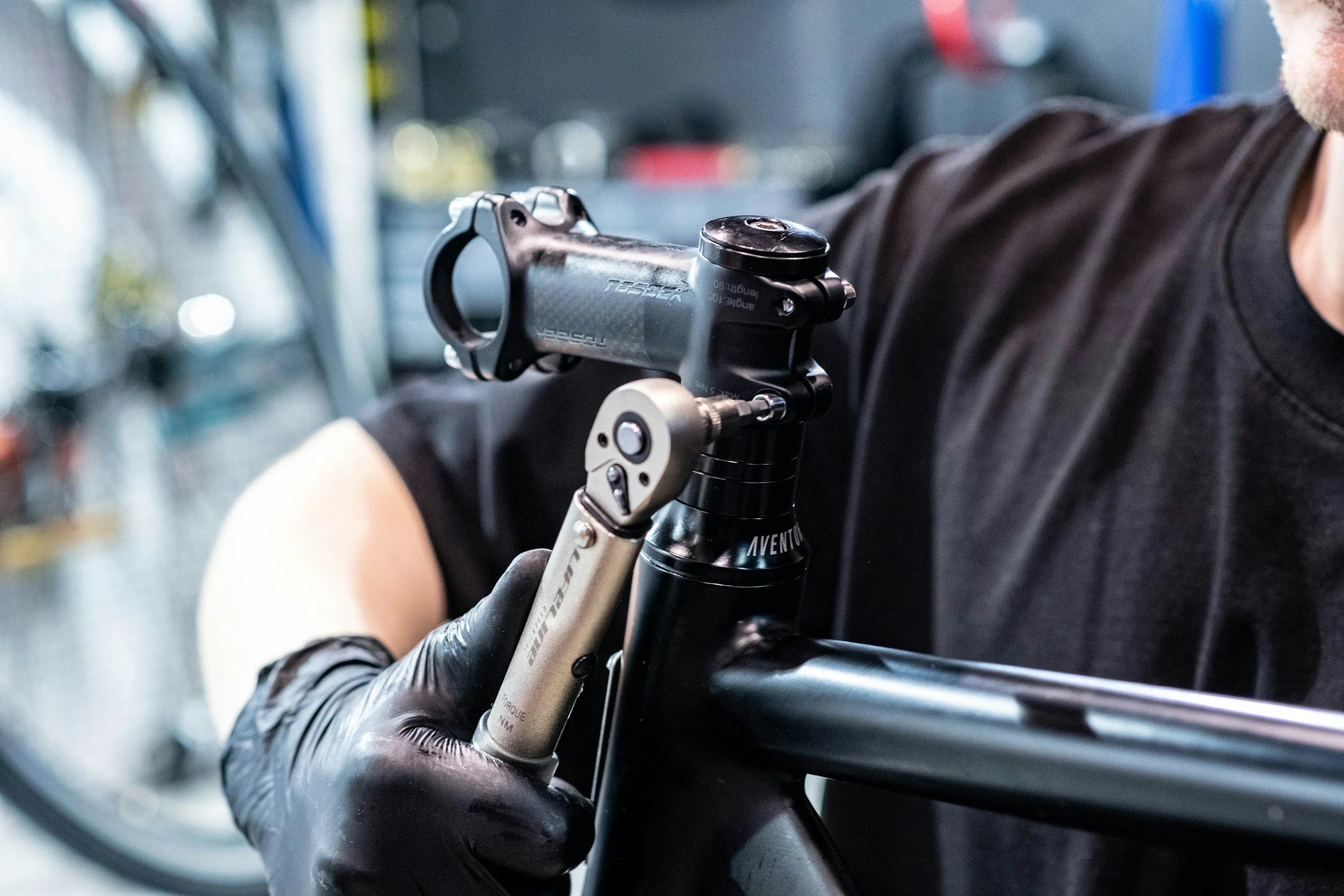 A person wearing black gloves is adjusting the suspension fork of a mountain bike in a workshop or bike repair shop.