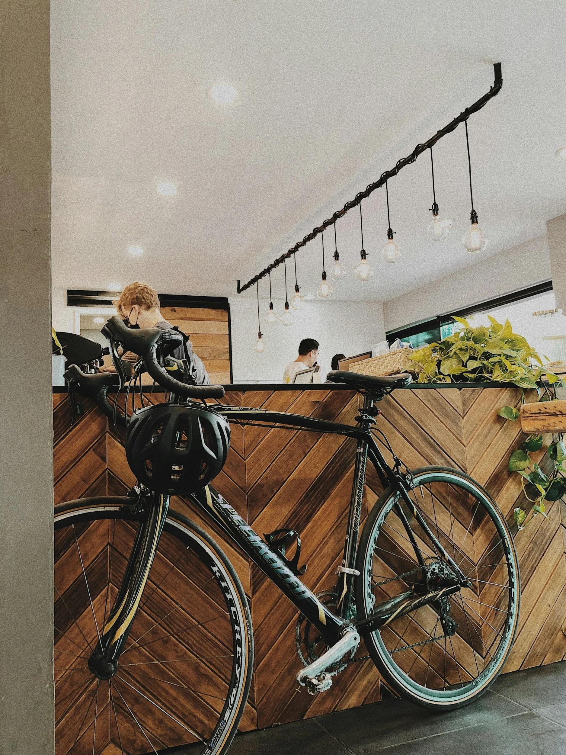 A black and teal road bicycle with a white saddle leaning against a wooden counter in a cafe, with people working on laptops in the background and industrial hanging light bulbs overhead.