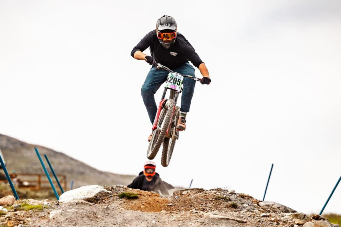 Stuart on a mountain bike wearing a helmet and goggles jumps off a rocky trail during a race, with another cyclist visible in the background.