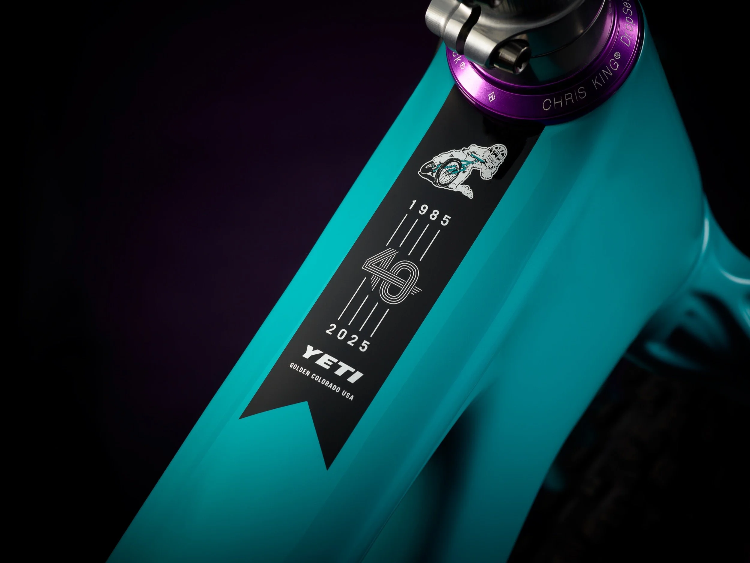 Close-up of a turquoise bicycle frame with a black sticker displaying a robotic figure riding a bicycle, the year 1985, and the years 2025, along with the brand name YETI and the location Golden, Colorado, USA. Part of the bicycle's purple and silver component is visible at the top.