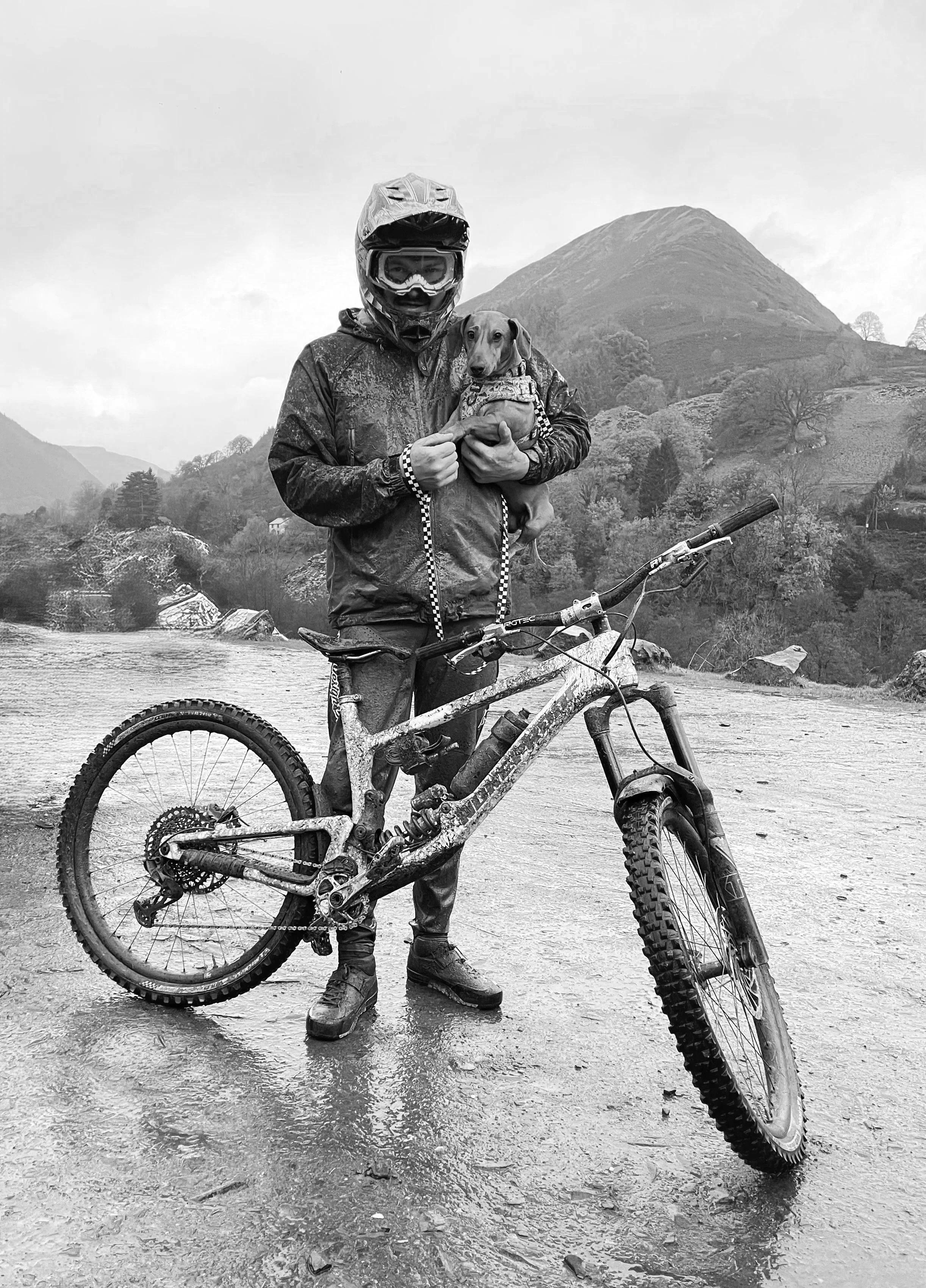 Person in motorcycle gear holding a dog on a mountain trail with mountains in the background.