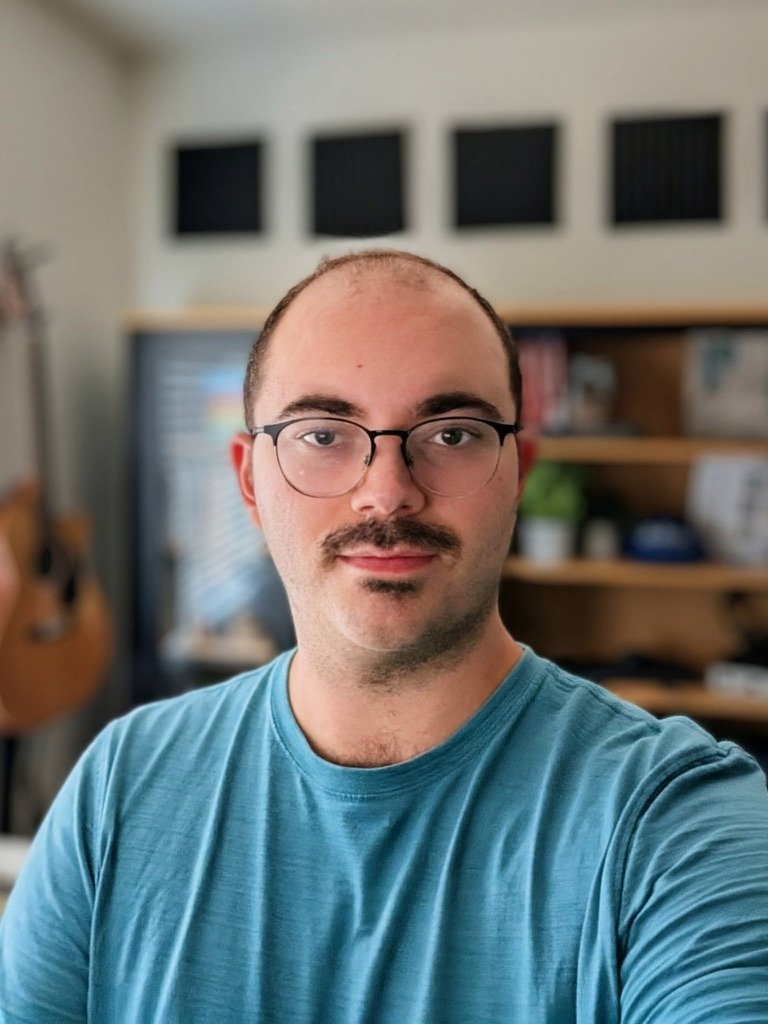 A man with glasses and a mustache taking a selfie in a room with various objects including a guitar and a bookshelf.