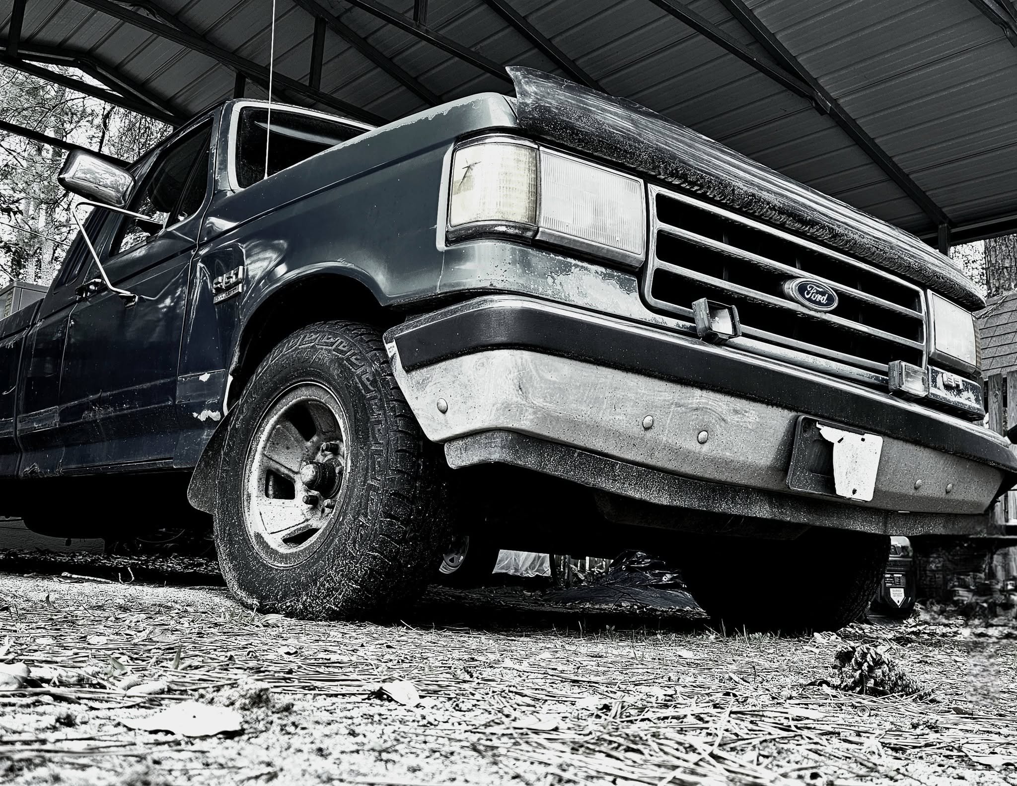 Front view of an old black Ford pickup truck parked under a metal carport, with dirt and leaves on the ground and trees in the background.