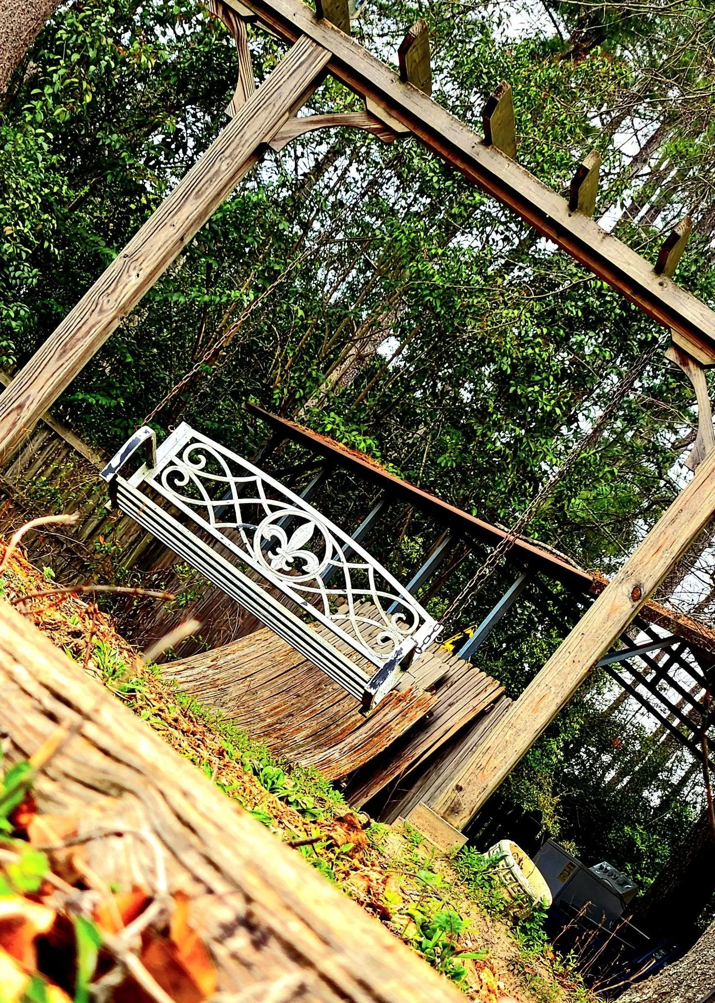 A white iron bench with decorative patterns is situated on a wooden deck in a backyard, surrounded by lush green trees and plants, and there are garden tools, a barbecue grill, and a black shed in the background.
