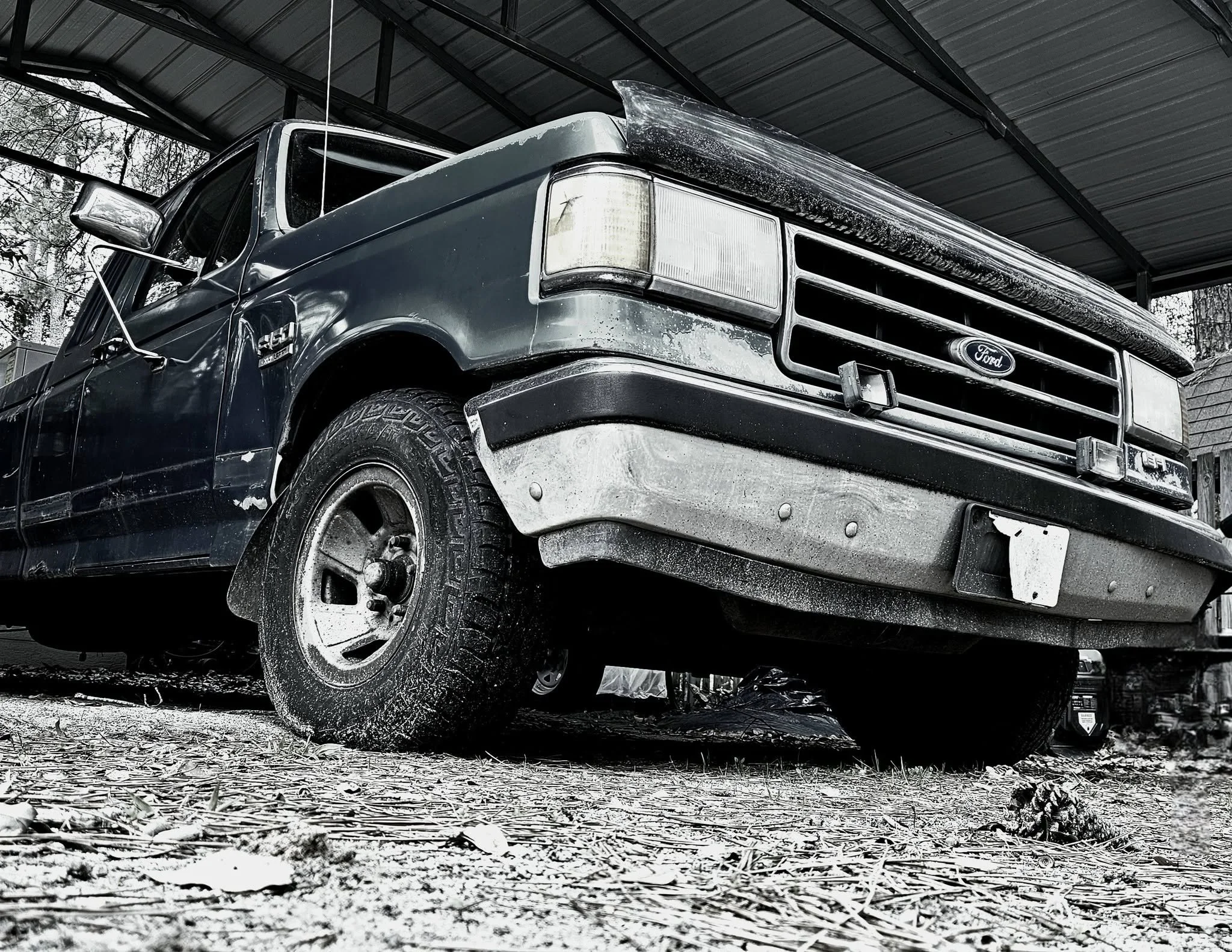Black pickup truck parked under a metal roof, with dirt and leaves on the ground, and trees in the background.