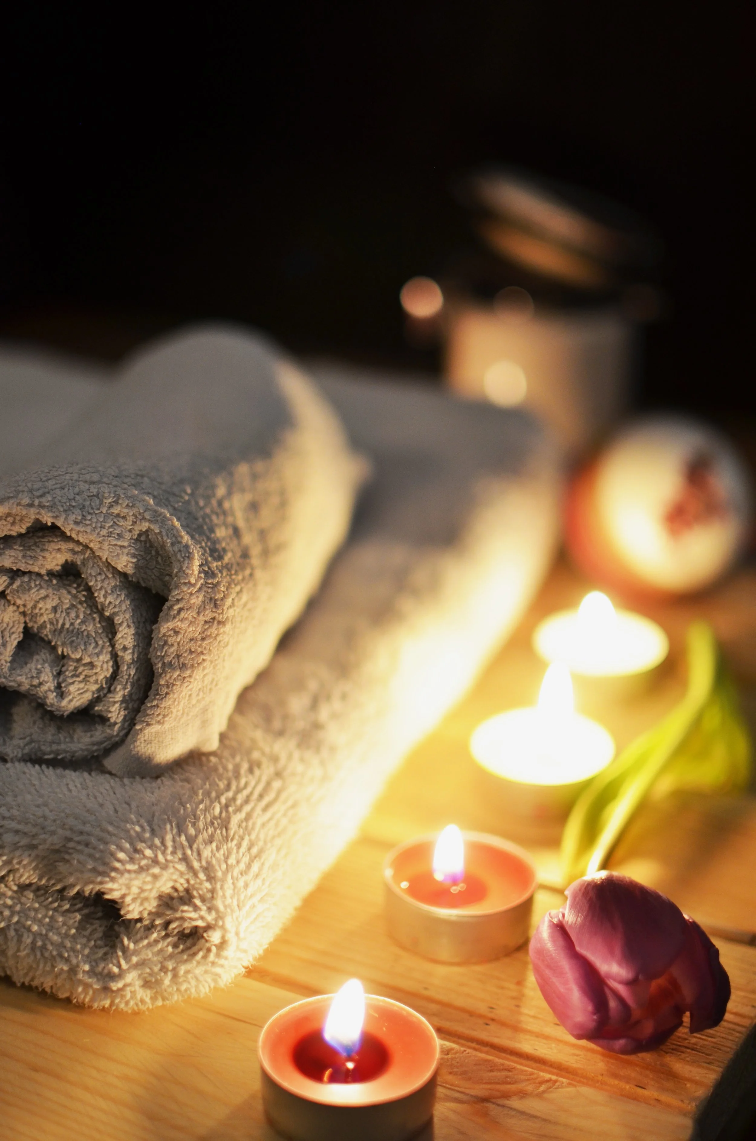 Close-up of a rolled grey towel, surrounded by candles, a pink rose, and spa items, creating a relaxing ambiance.