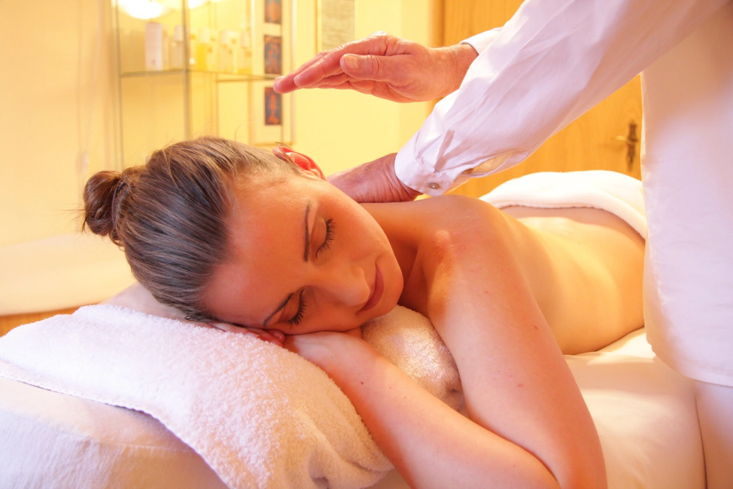 A woman receiving a massage in a spa, lying face down on a massage table with a towel over her lower back, woman looks relaxed with eyes closed, massage therapist wearing white uniform, soft warm lighting, relaxing spa decor in background.