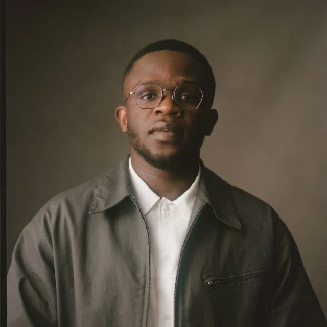 A young man with glasses and a beard wearing a white shirt and a dark jacket against a plain background.