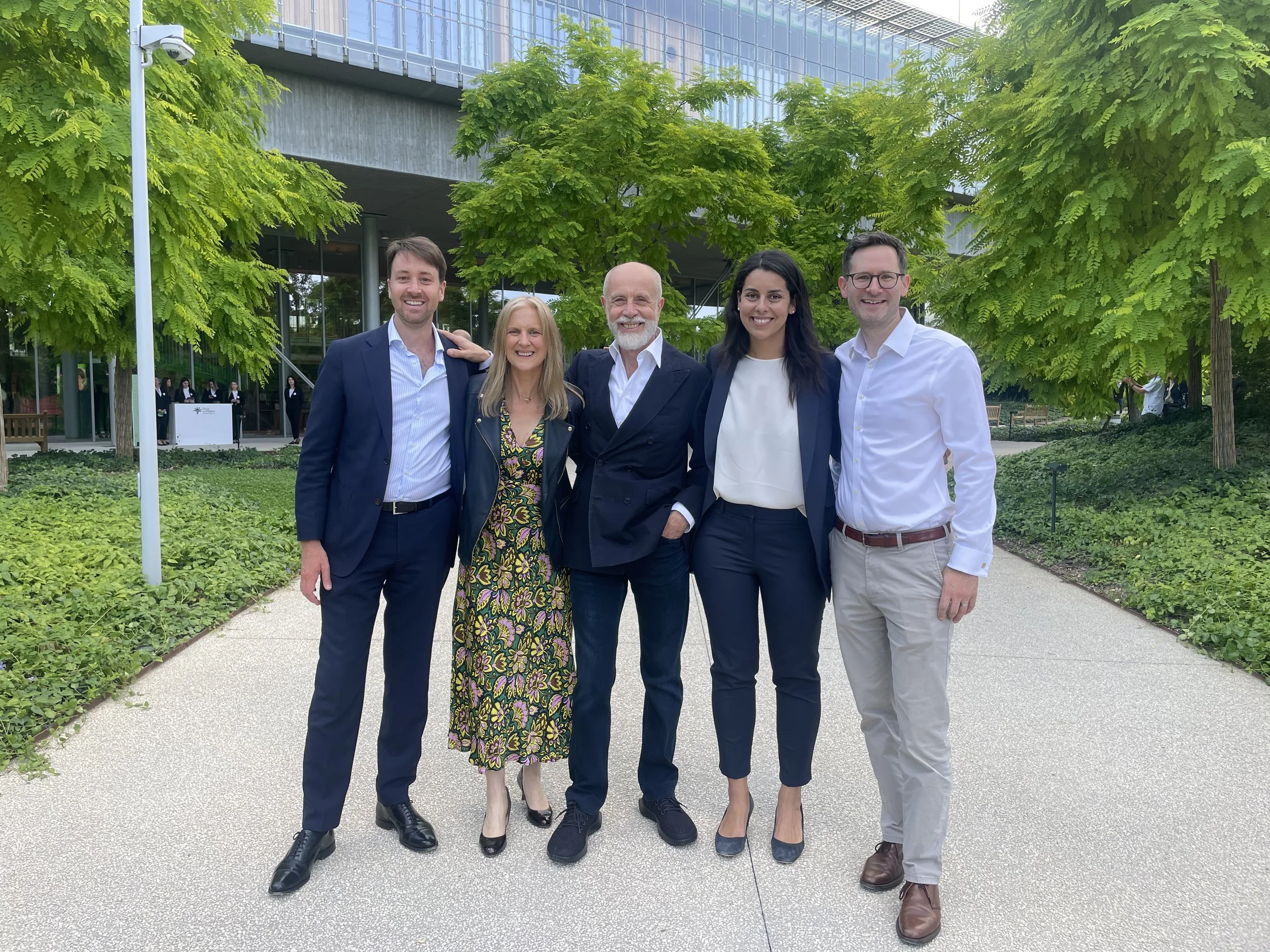 Five smiling individuals in front of a lush green grounds with sleek building.