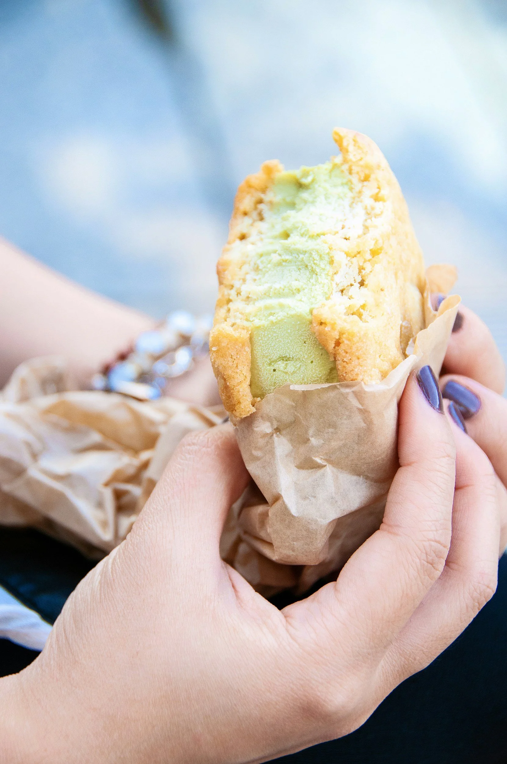 Close-up of a person holding a partially eaten ice cream cone with vanilla and green ice cream, wrapped in brown paper, with a blue background.